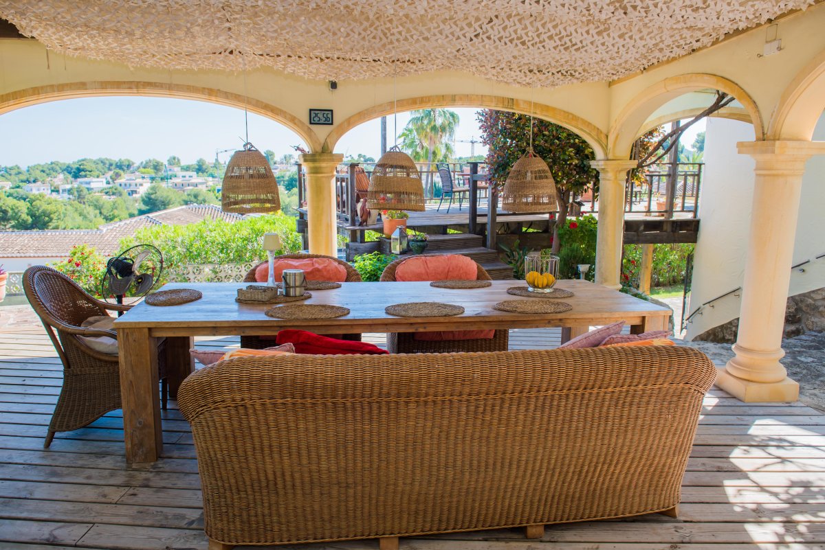 Porche con mesa de madera rústica y asientos de mimbre, iluminado con lámparas colgantes de mimbre, con vistas al paisaje.