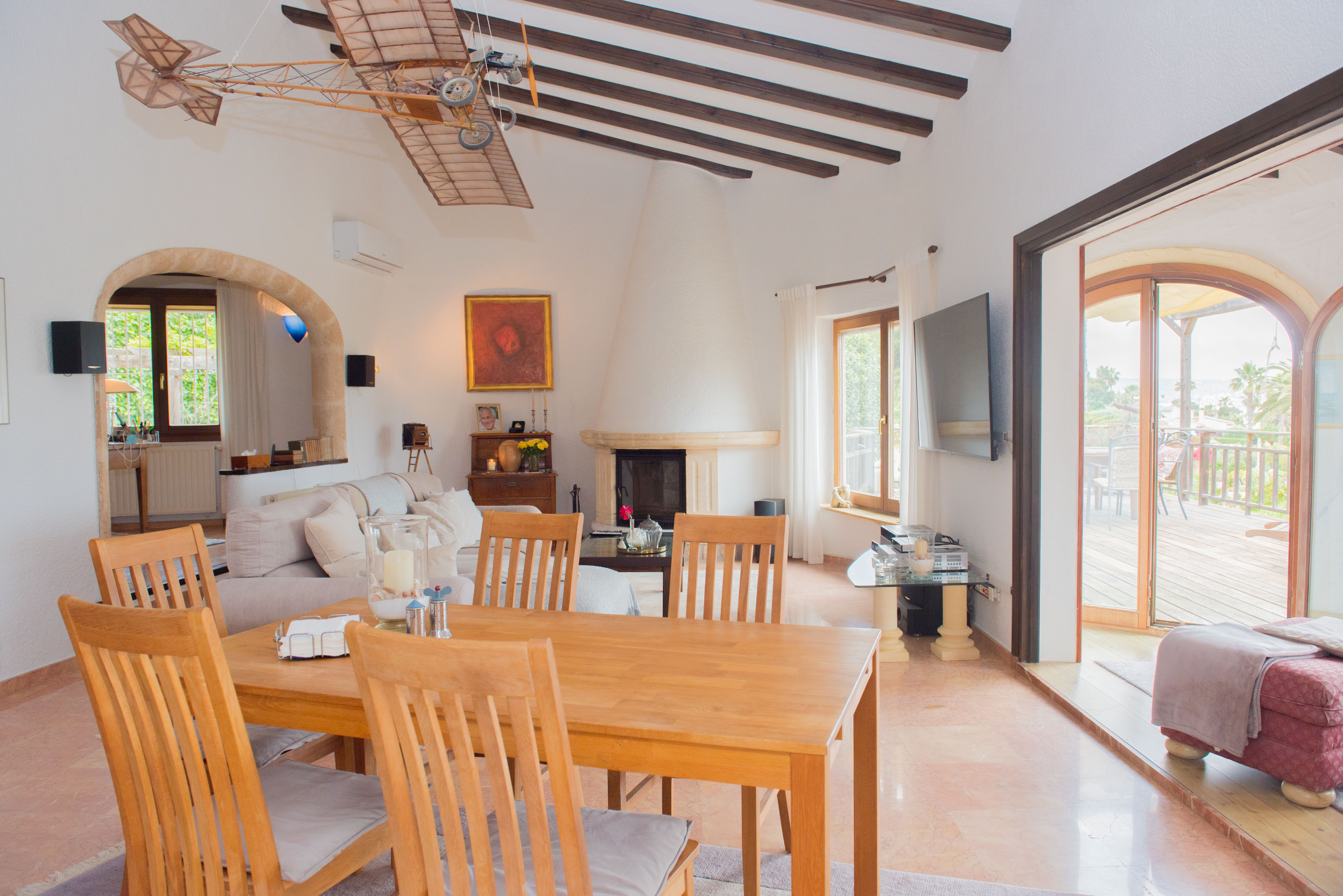 Salón comedor con vigas de madera, chimenea cónica blanca y acceso directo a la terraza de madera a través de grandes ventanales arqueados.