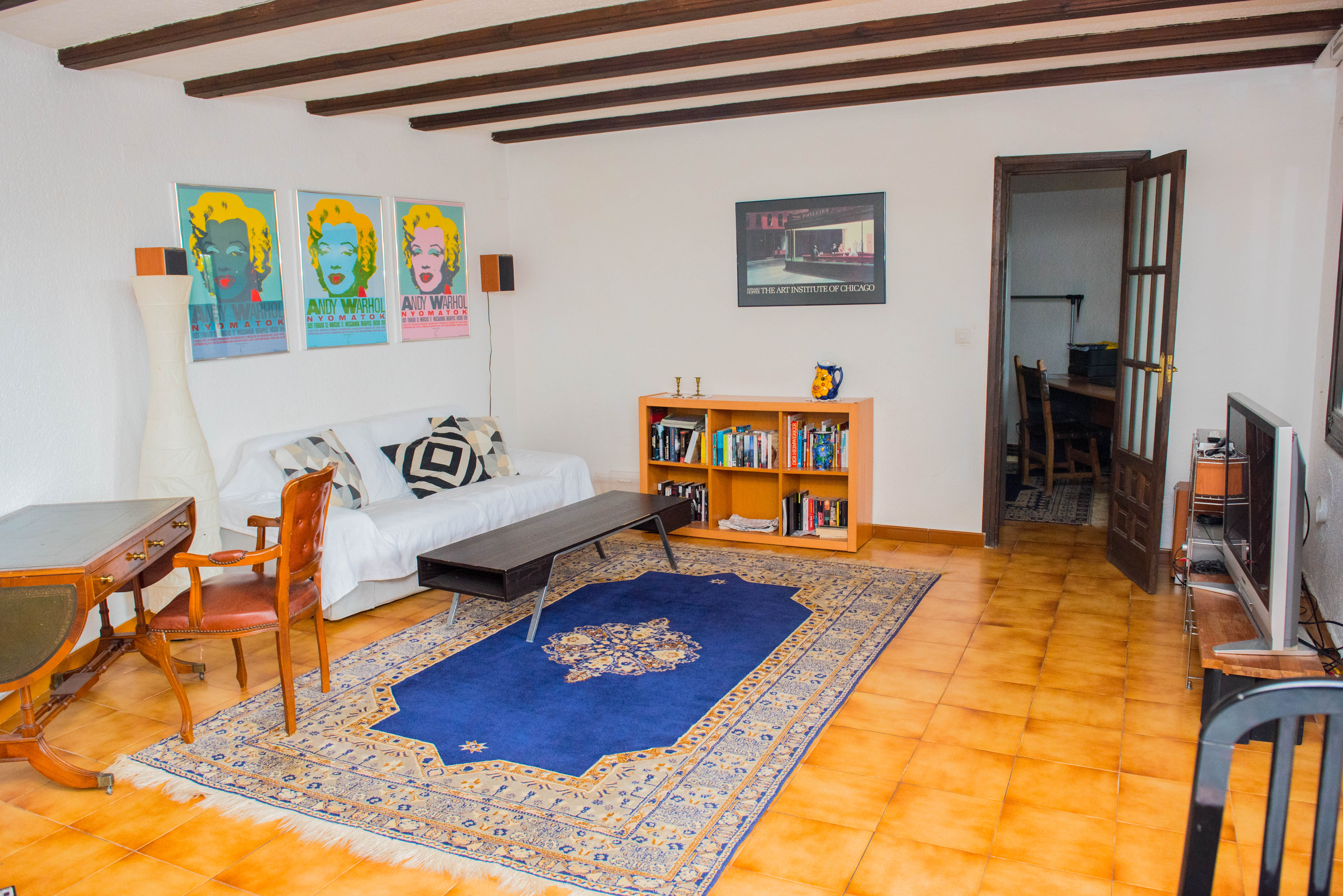 Salón con vigas de madera a la vista, suelo de baldosas cerámicas, zona de estar con sofá blanco, escritorio clásico y acceso a un despacho contiguo.