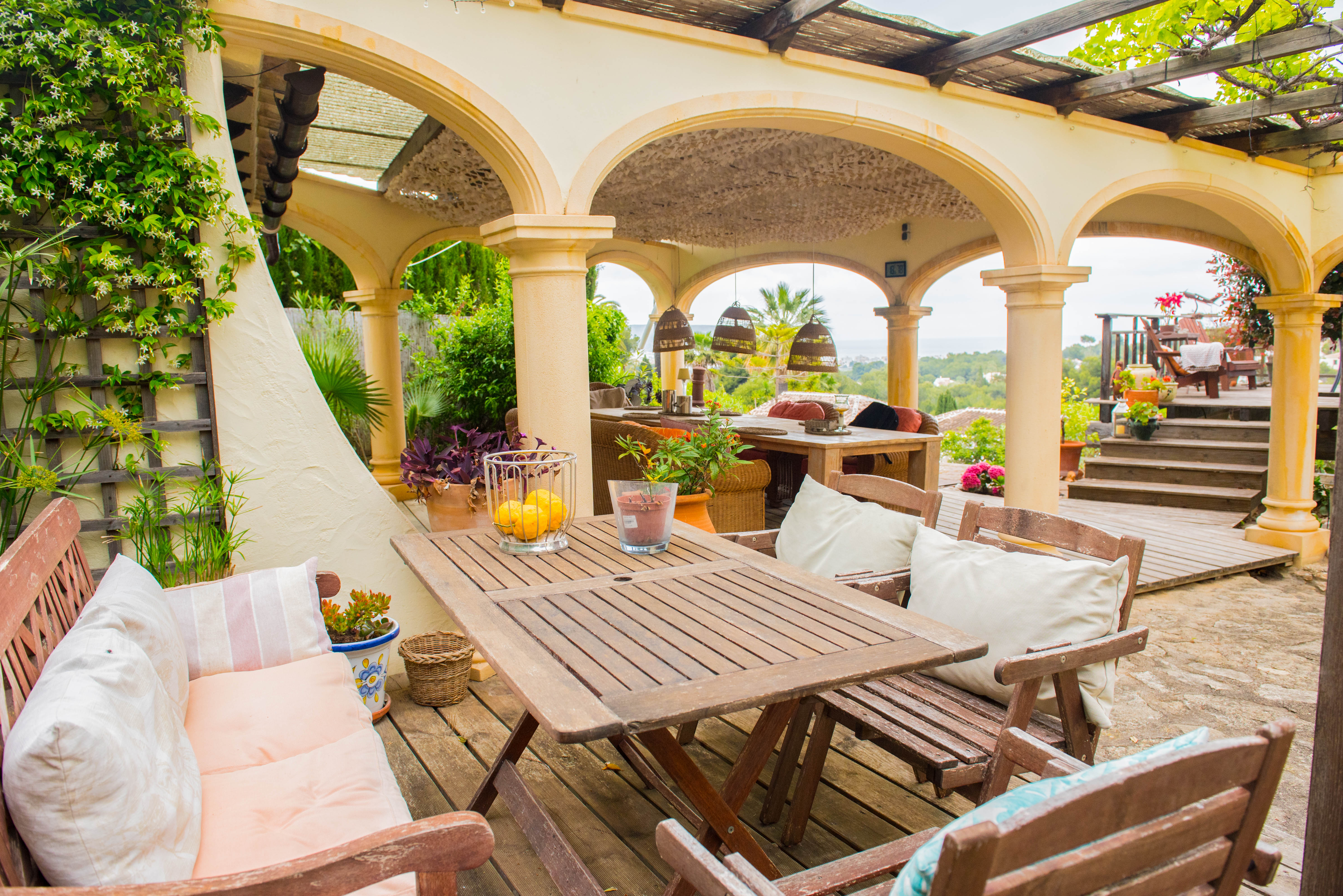 Terraza cubierta con arcos mediterráneos, mobiliario de comedor en madera, suelos de tarima y vistas despejadas al mar y al entorno natural.