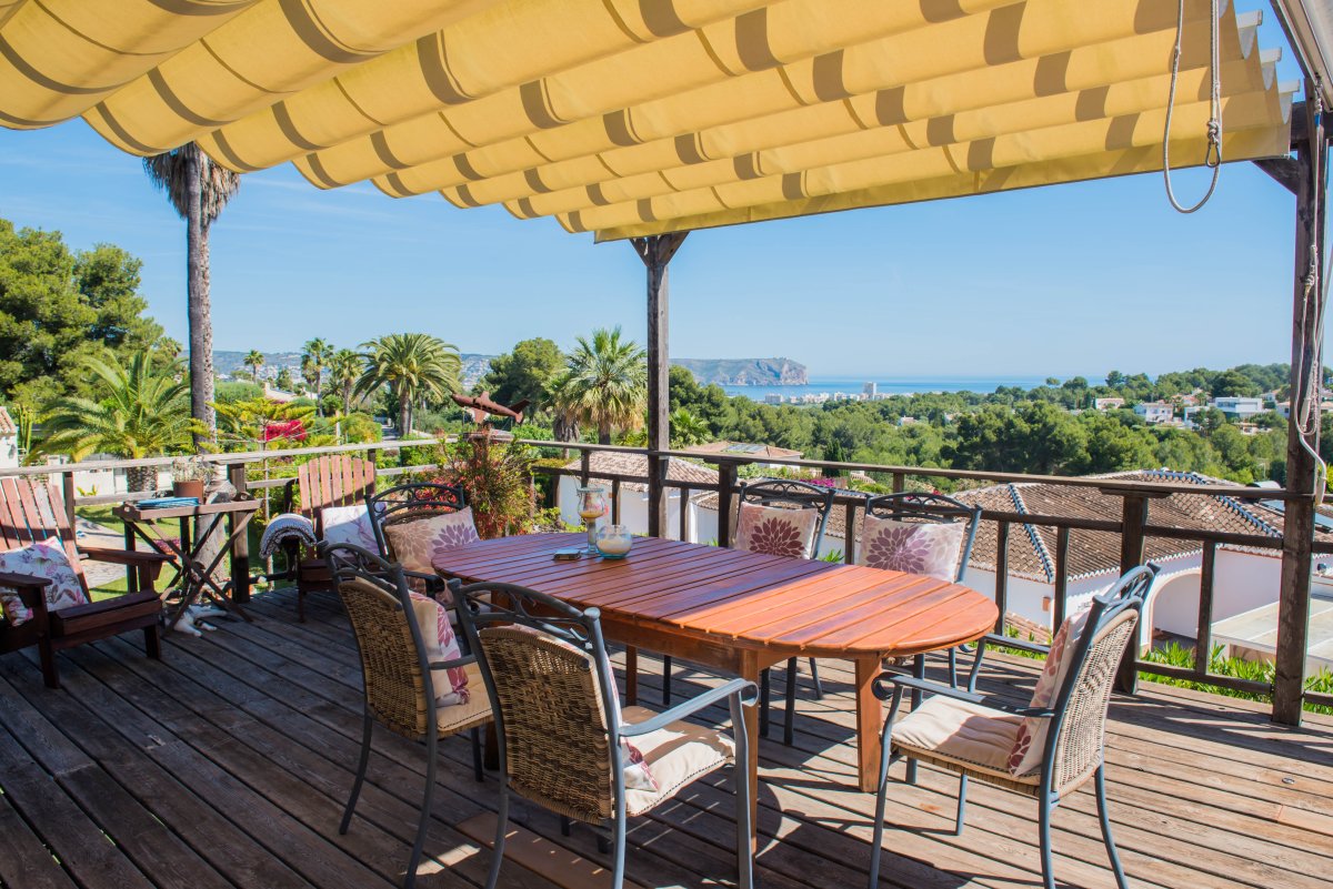 Terraza de madera con toldo y mesa de comedor con vistas al mar y vegetación.