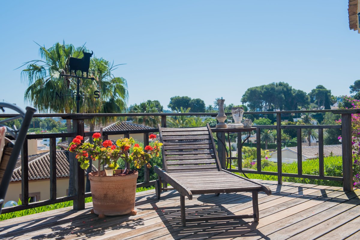 Terraza de madera con tumbona, vistas al jardín y detalles decorativos como un veleta de toro y macetas de geranios.