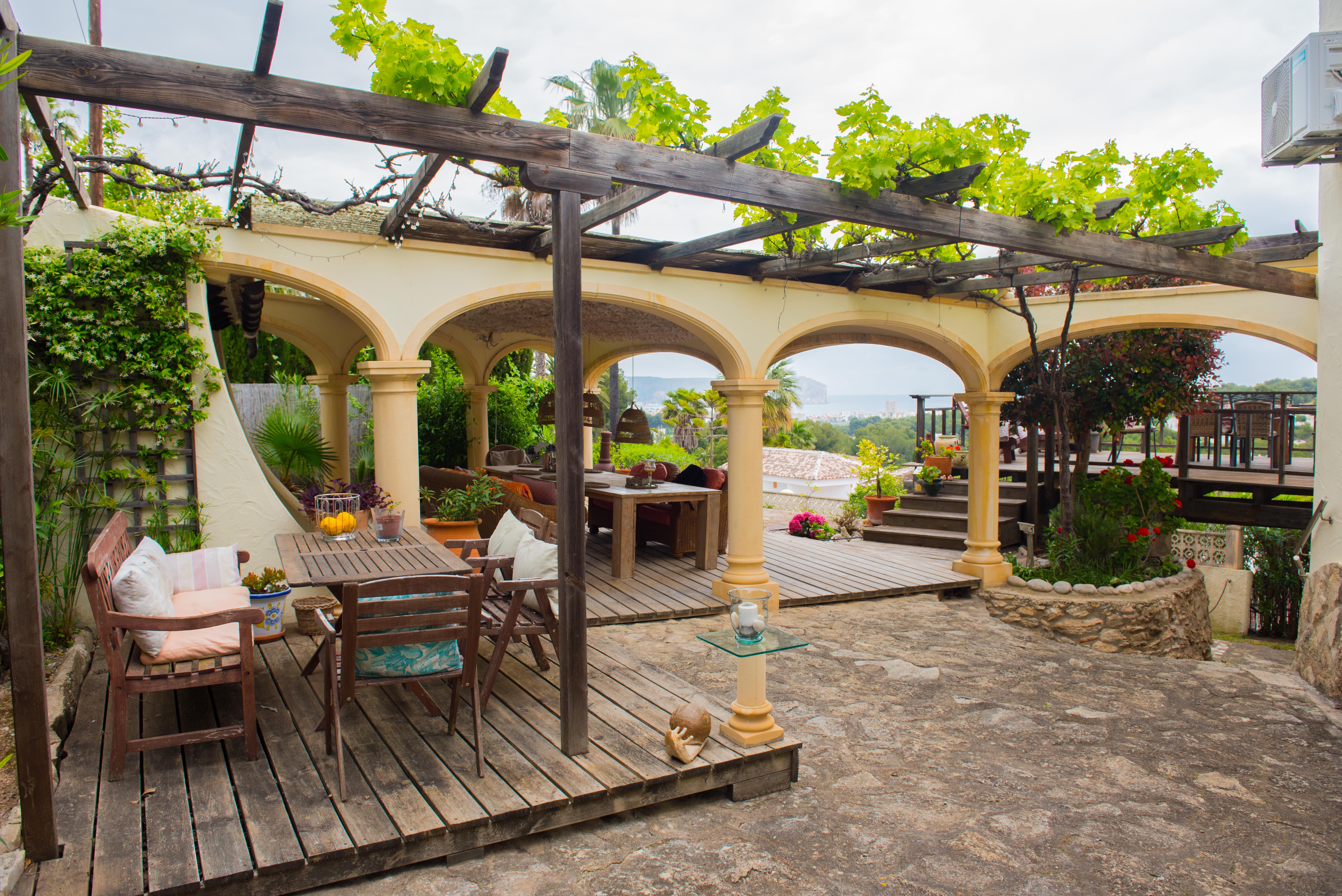 Terraza exterior con pérgola de madera y vides, zona de estar sobre tarima y porche de arcos con vistas panorámicas al mar y al entorno costero.