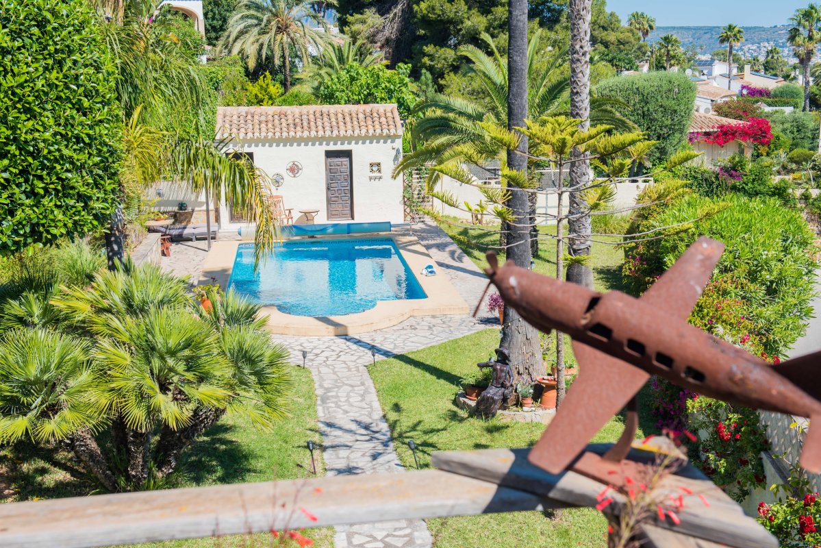 Vista de la piscina con borde de piedra, caseta con tejas y jardín tropical desde una terraza con decoración de avión oxidado.