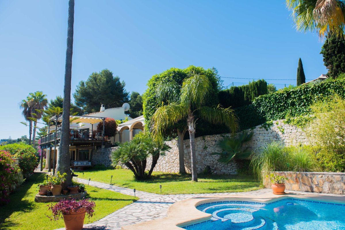 Vista del jardín con piscina de mosaico azul y camino de piedra que conduce a la villa de estilo mediterráneo.