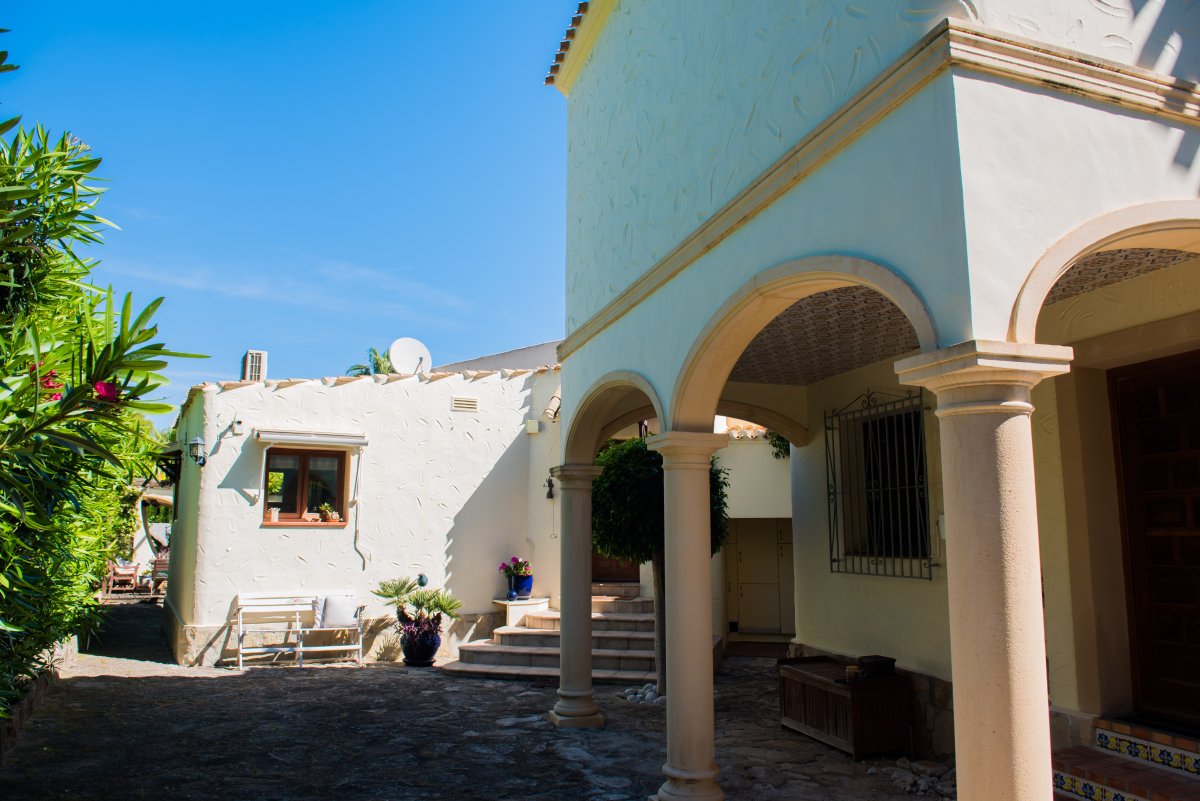 Vista exterior de la villa con arcos de piedra y paredes texturizadas blancas, rodeada de vegetación mediterránea.