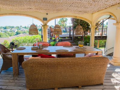Porche con mesa de madera rústica y asientos de mimbre, iluminado con lámparas colgantes de mimbre, con vistas al paisaje.