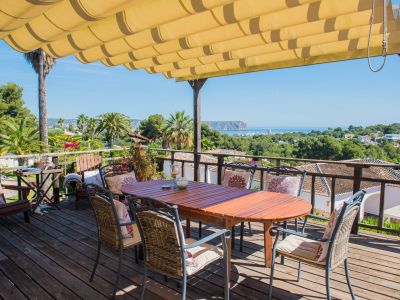Terraza de madera con toldo y mesa de comedor con vistas al mar y vegetación.