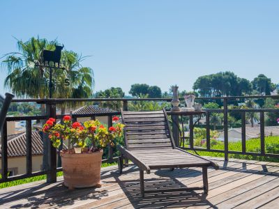 Terraza de madera con tumbona, vistas al jardín y detalles decorativos como un veleta de toro y macetas de geranios.