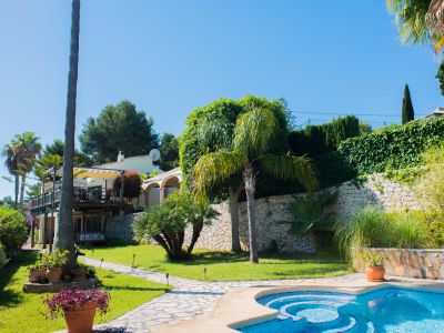 Vista del jardín con piscina de mosaico azul y camino de piedra que conduce a la villa de estilo mediterráneo.