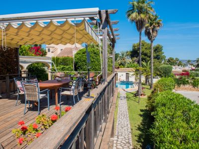 Vista desde la terraza de madera con toldo, mesa y sillas hacia la piscina y jardines.