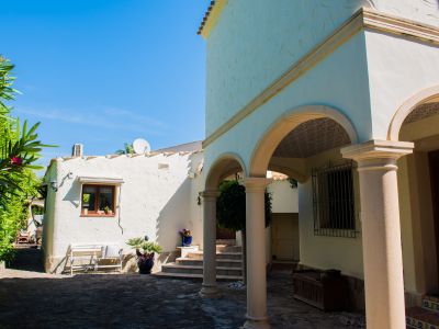 Vista exterior de la villa con arcos de piedra y paredes texturizadas blancas, rodeada de vegetación mediterránea.