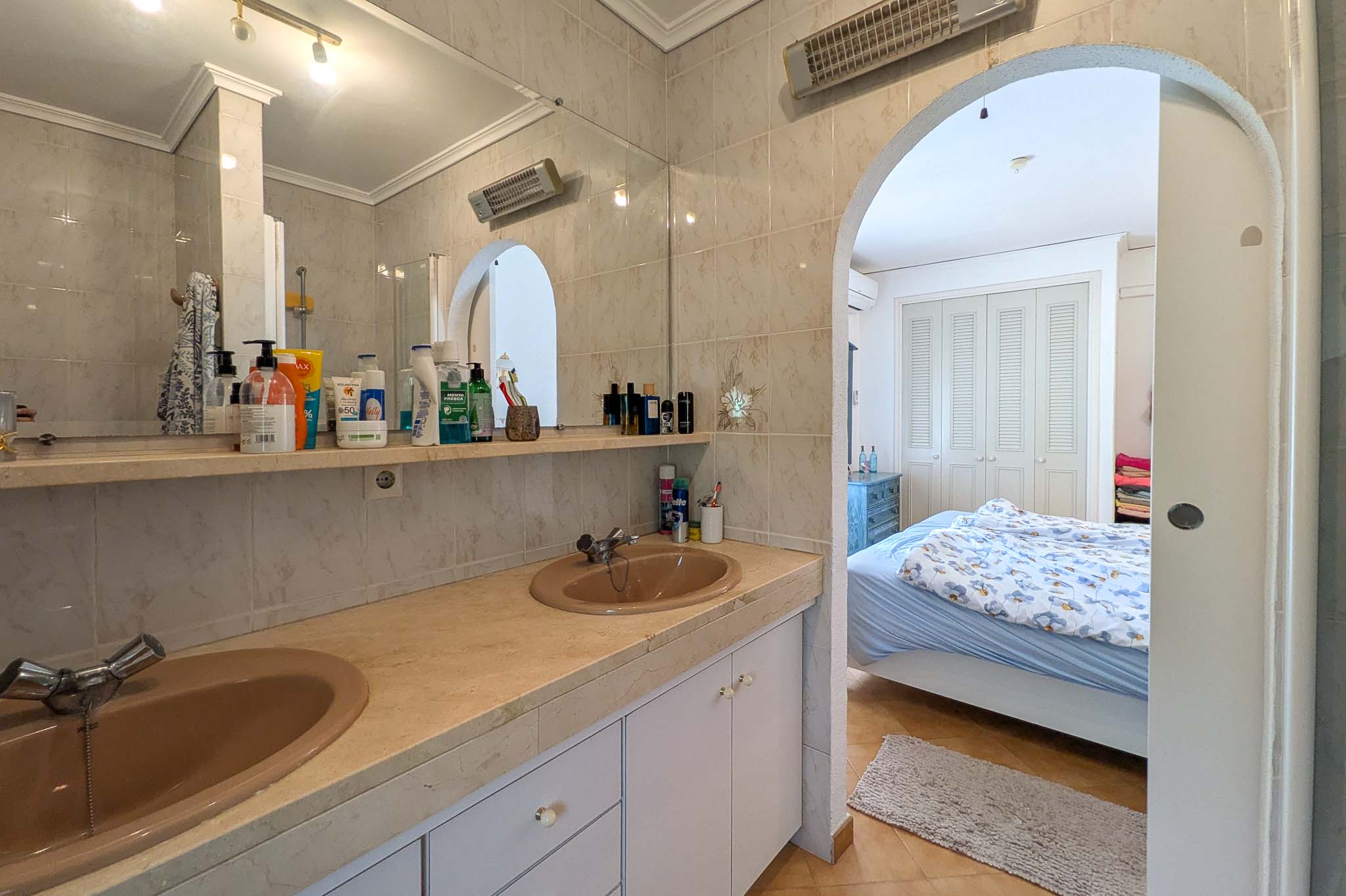 Baño en suite con lavabo de doble seno sobre encimera de mármol, conectado al dormitorio principal mediante un arco arquitectónico.