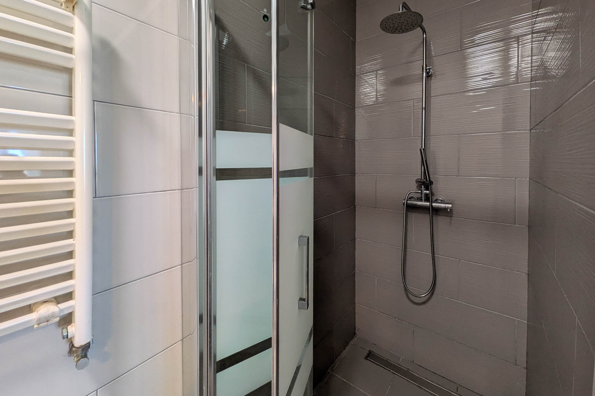 Baño funcional con ducha a ras de suelo, mampara de cristal con bandas traslúcidas, grifería cromada y radiador toallero blanco sobre revestimiento cerámico.