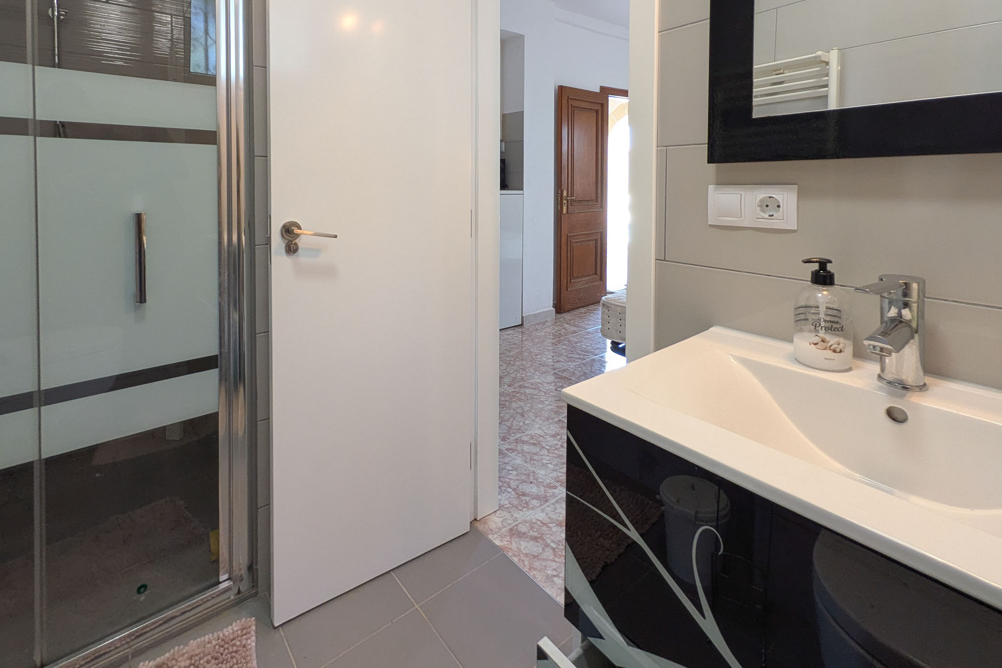 Baño funcional con mueble de lavabo negro decorado, mampara de ducha de cristal y suelos cerámicos grises. La estancia conecta con un pasillo de baldosas tradicionales.