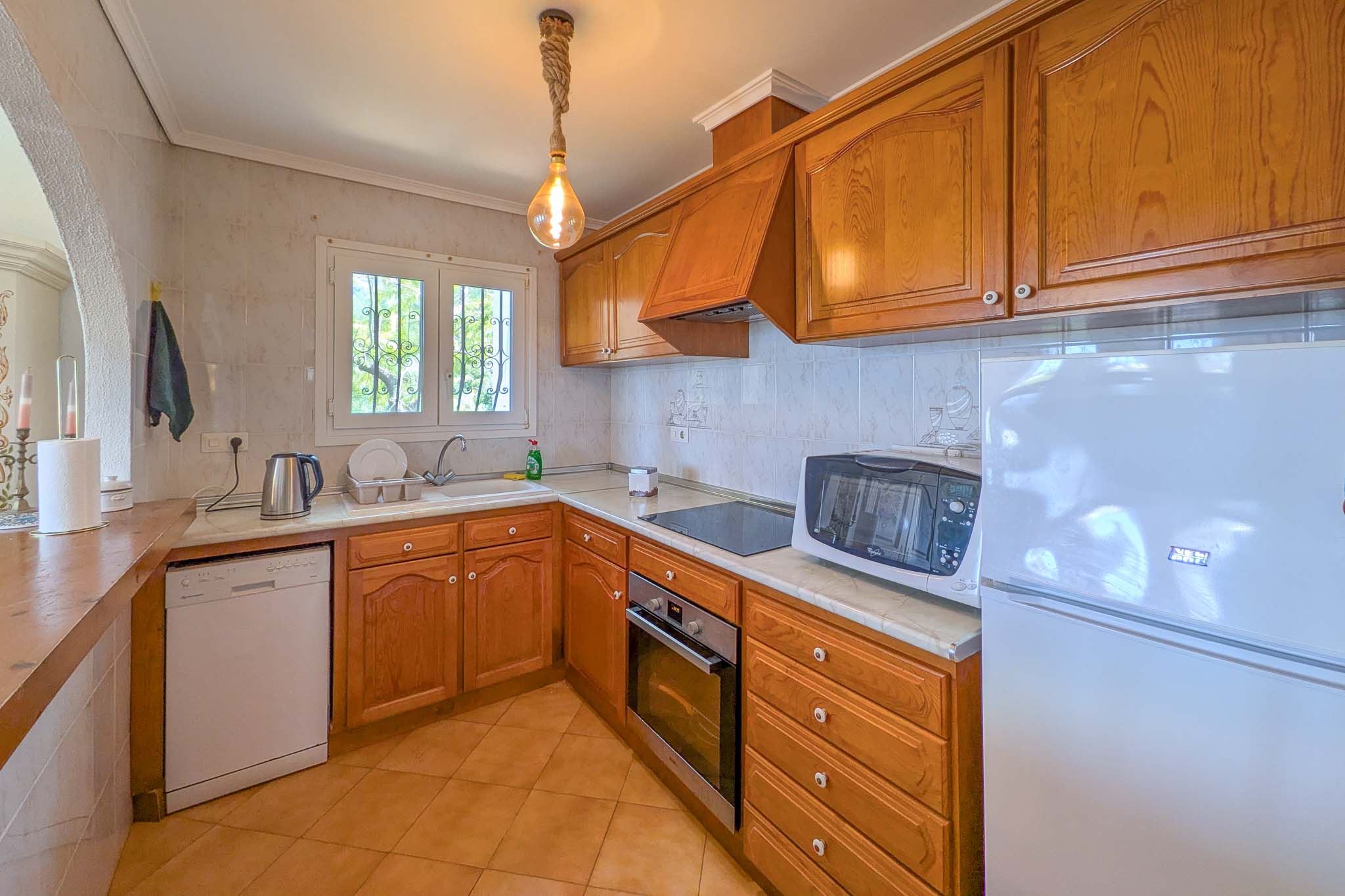 Cocina de estilo rústico con armarios de madera, encimera de mármol y electrodomésticos integrados. Destaca su iluminación con lámpara de cuerda y el acceso de luz natural.