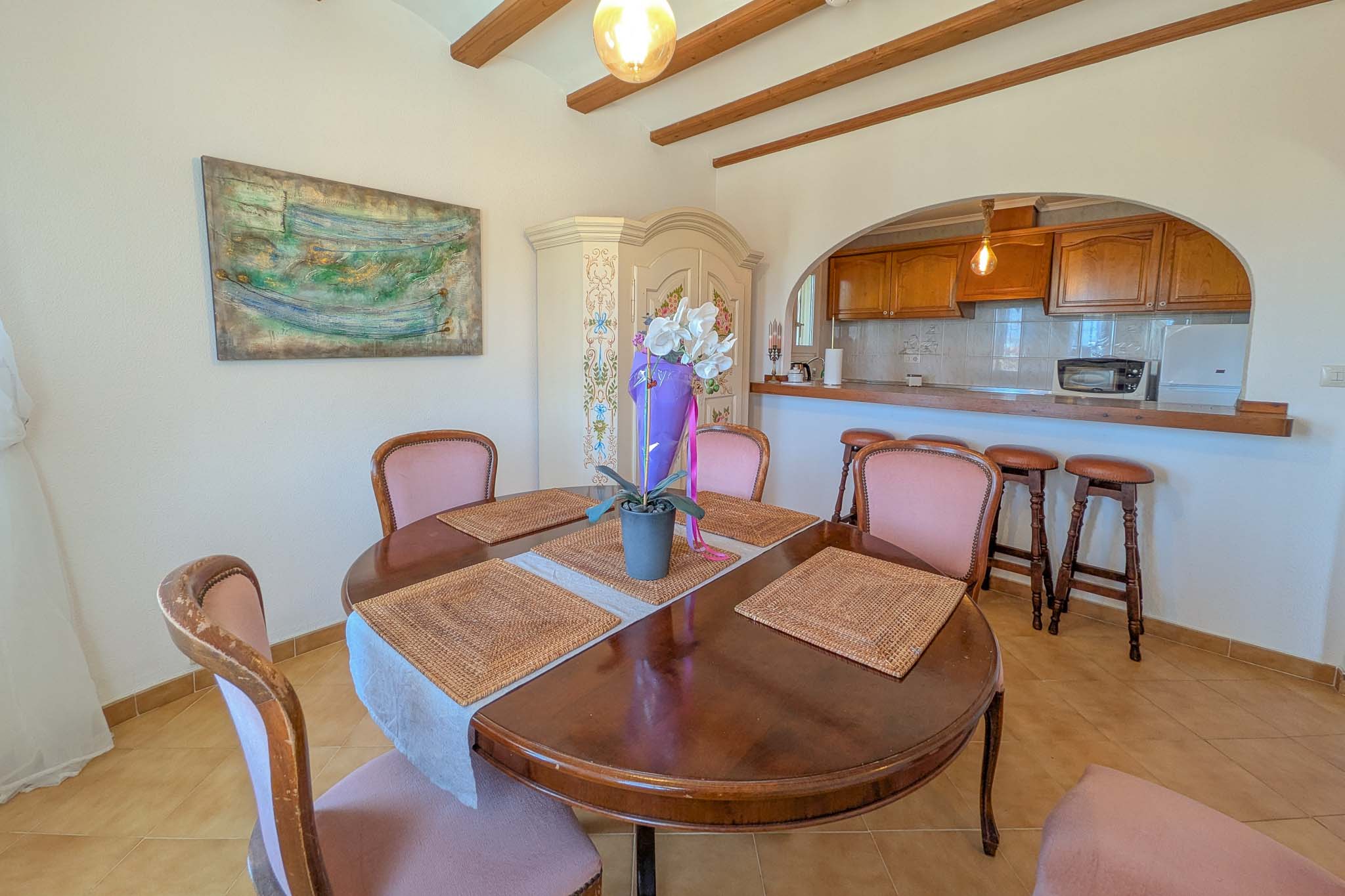 Comedor con mesa circular de madera y vigas vistas, conectado a la cocina semiabierta mediante un arco con barra de desayuno y taburetes rústicos.