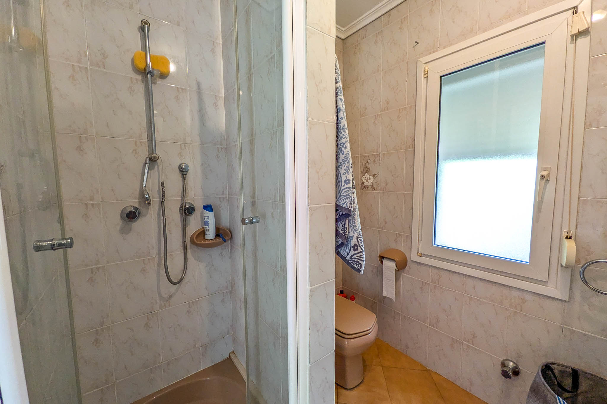 Cuarto de baño con ducha de mampara acristalada, inodoro en tonos crema y ventana de PVC blanco que aporta luz natural. Paredes revestidas con azulejos cerámicos decorados.