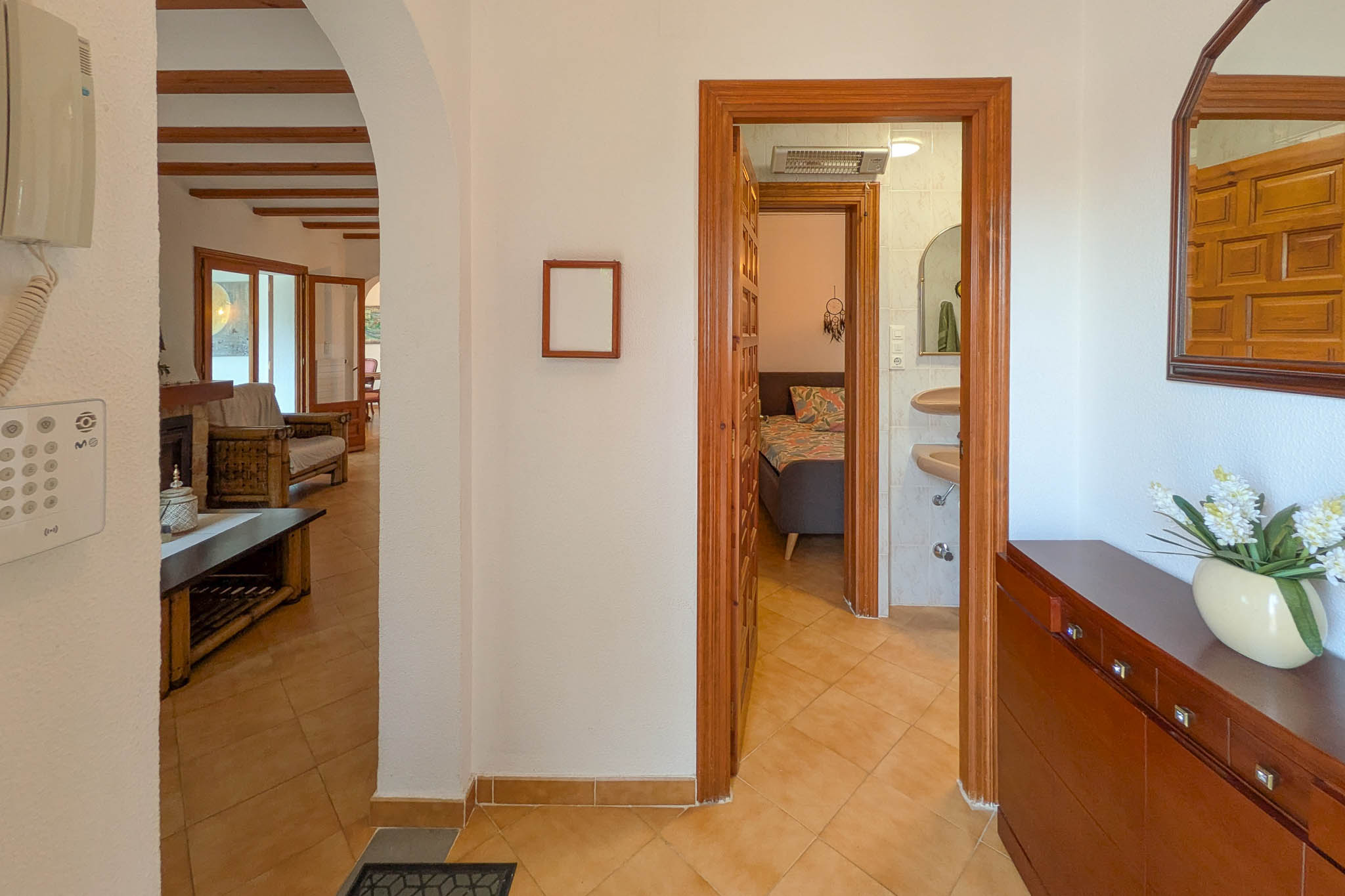 Distribuidor con suelos de gres cerámico que conecta el salón de vigas vistas con un dormitorio y baño, destacando la carpintería de madera natural y arcos tradicionales.