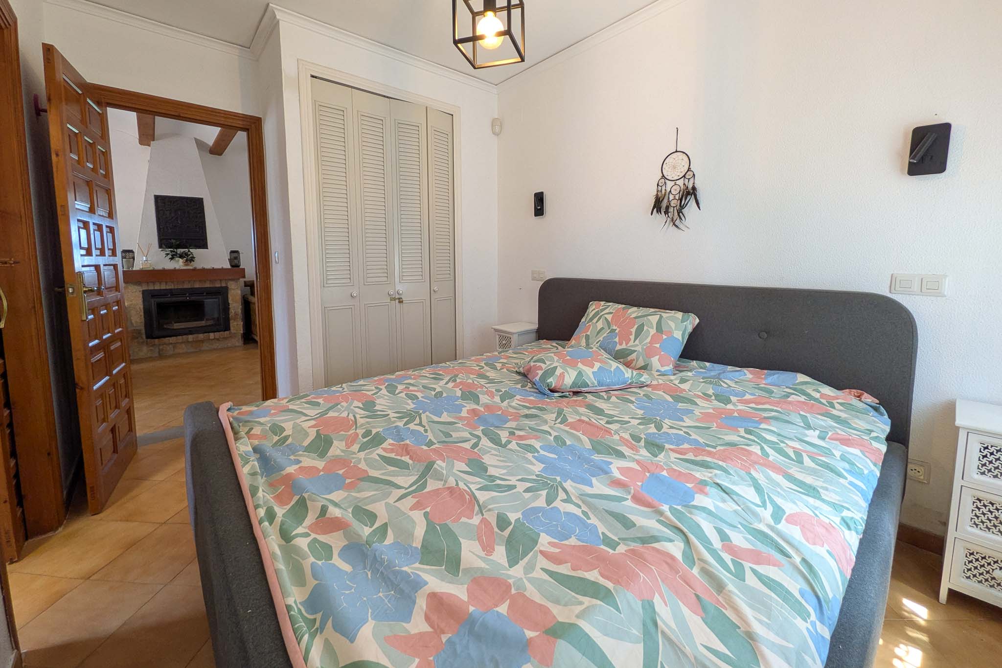 Dormitorio con armario empotrado de puertas mallorquinas blancas y vista directa al salón con chimenea a través de una puerta de madera maciza.