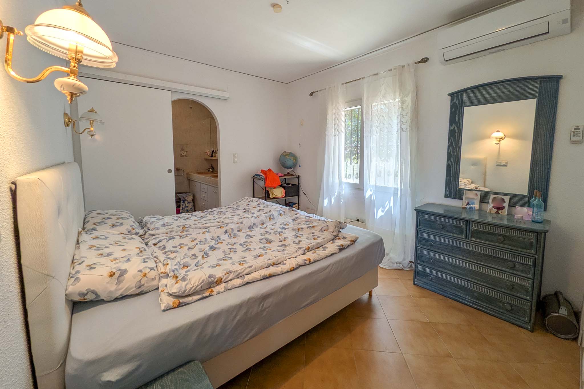 Dormitorio con baño en suite, aire acondicionado tipo split, suelos de gres y mobiliario de madera. La estancia cuenta con iluminación natural y acceso directo al baño.