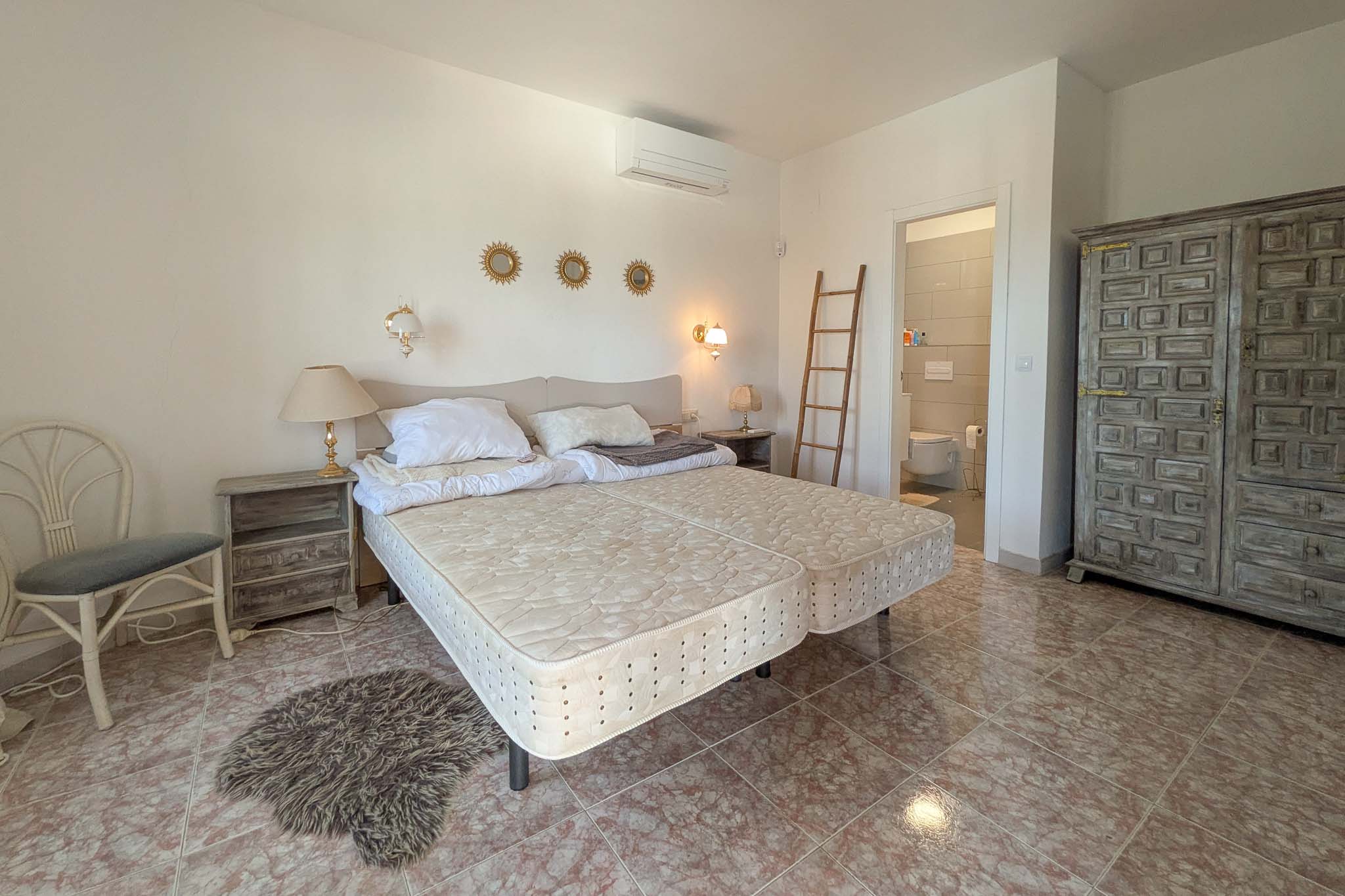 Dormitorio con baño en suite, suelos de gres efecto mármol, armario de madera tallada de estilo rústico y unidad de aire acondicionado instalada.