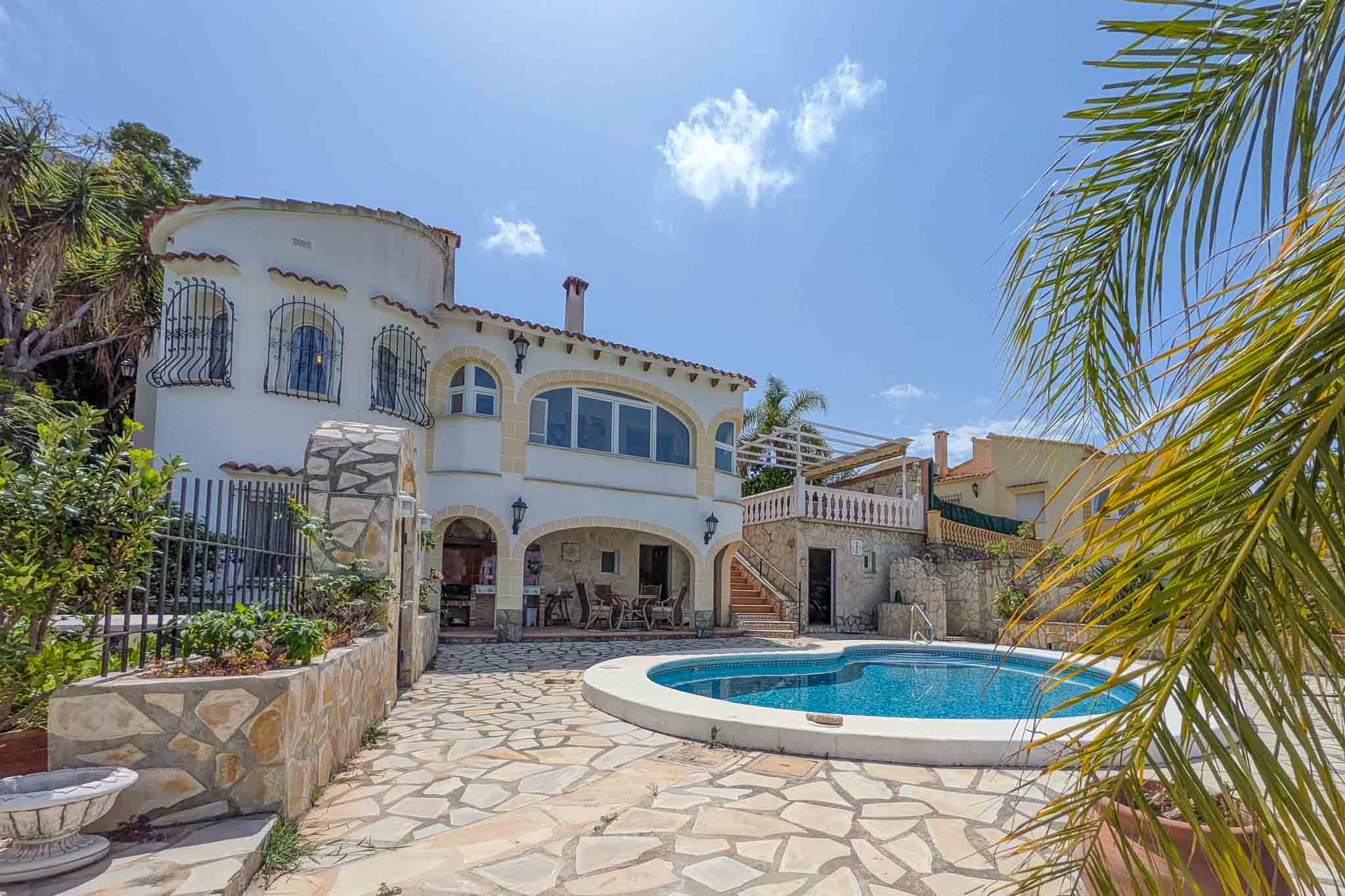 Exterior de la villa con piscina curva, terraza de piedra natural y porche con cocina de verano. La fachada blanca integra arcos tradicionales y una torre lateral.