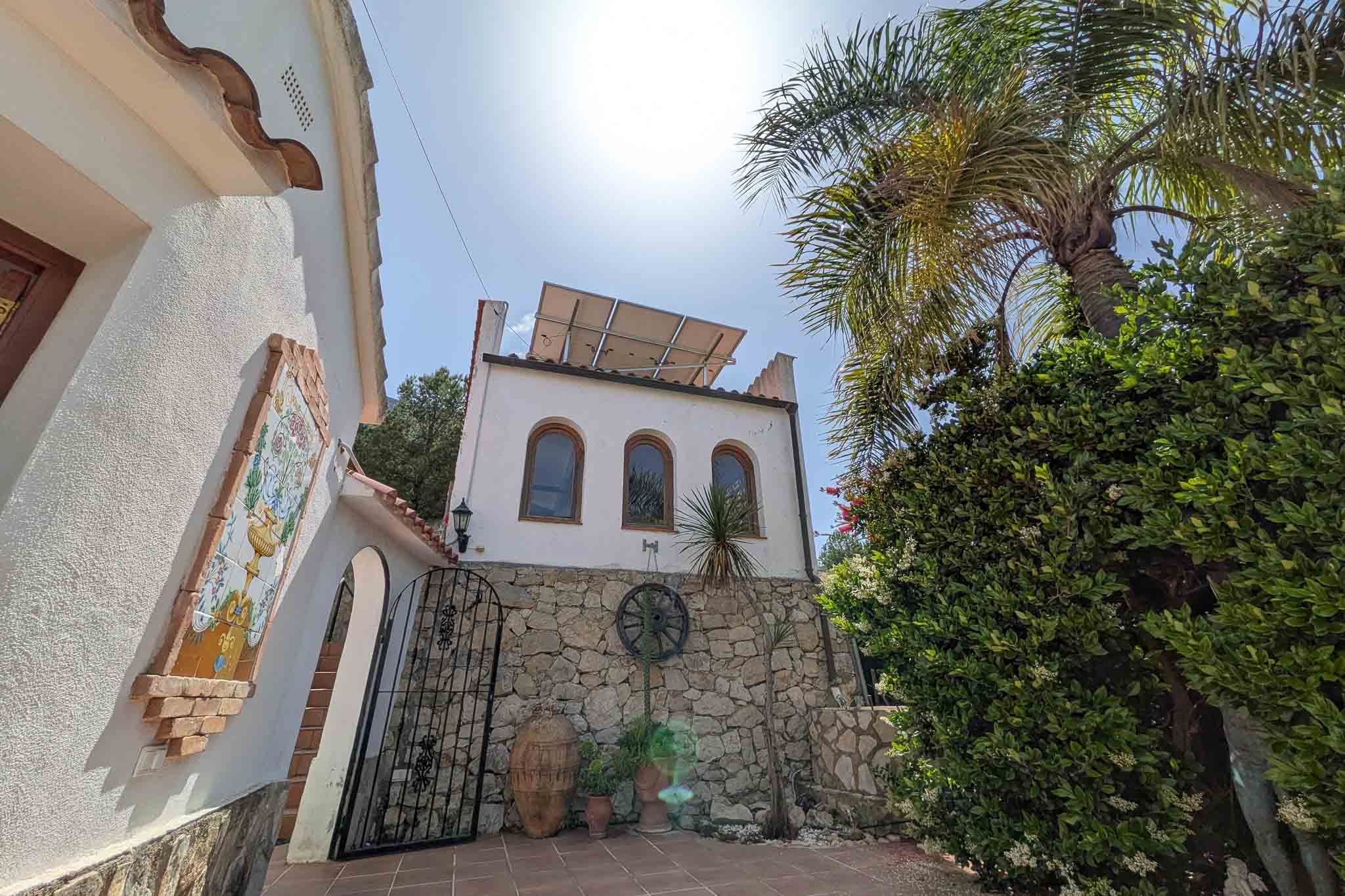 Fachada mediterránea con muros de piedra natural, paneles solares en el tejado y detalles tradicionales como un mural cerámico y una reja de forja.