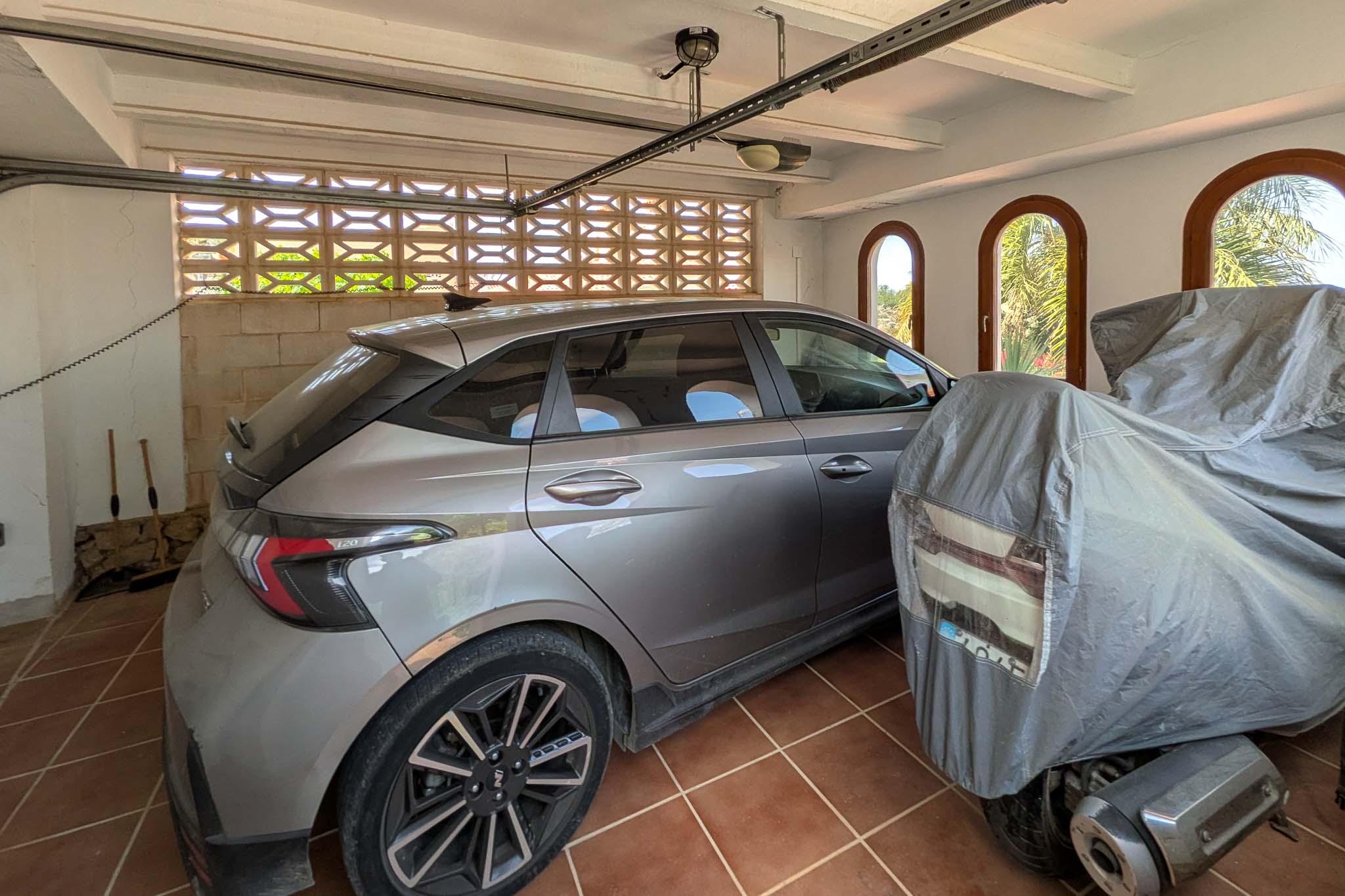 Garaje cubierto con suelo de gres, techo de vigas blancas y ventanas de arco. Incluye un muro de celosía para ventilación natural y espacio para coche y moto.