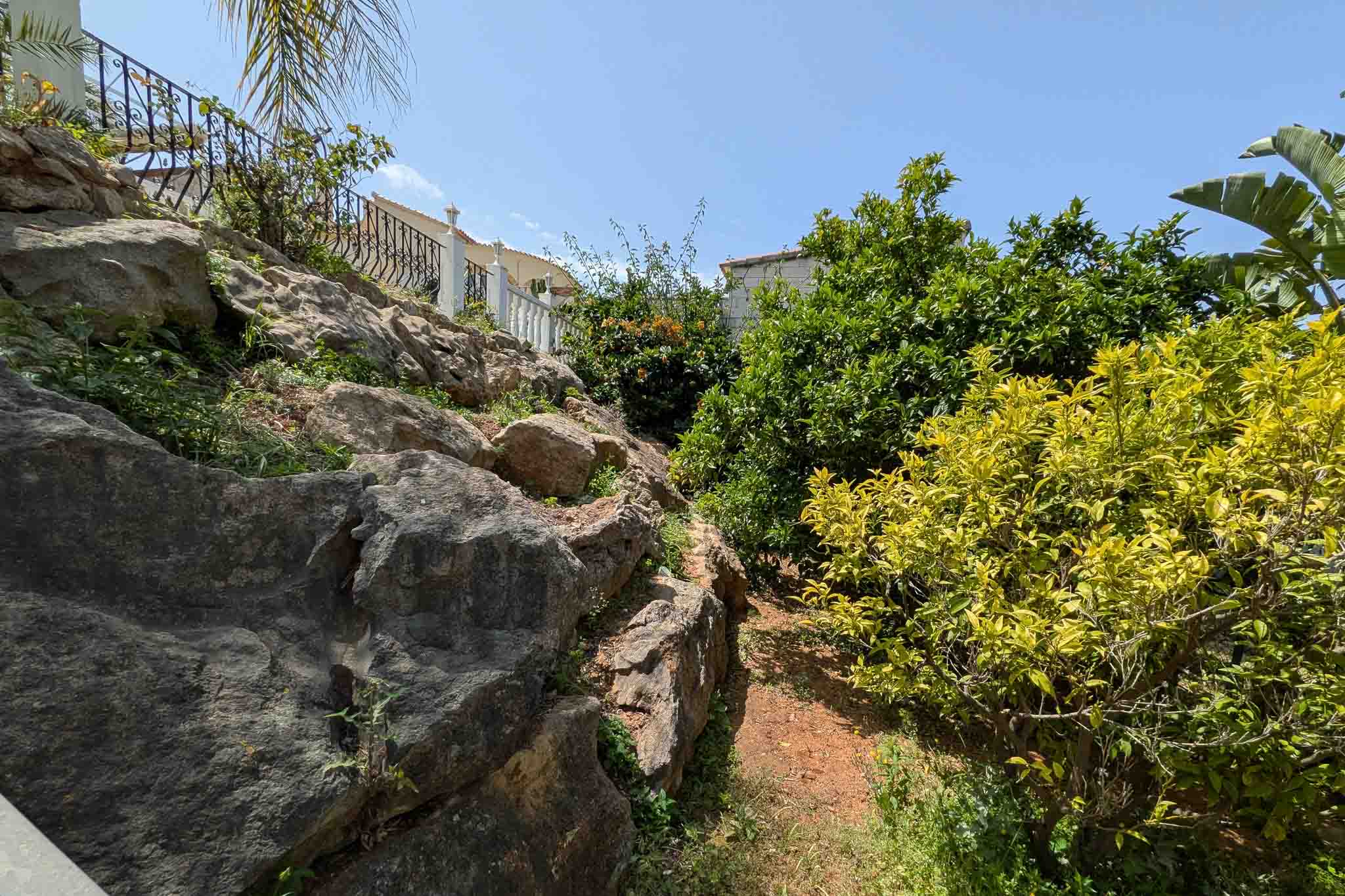 Jardín en cascada con formaciones de roca natural, senderos de tierra y vegetación mediterránea, incluyendo cítricos bajo el sol de La Pedrera.