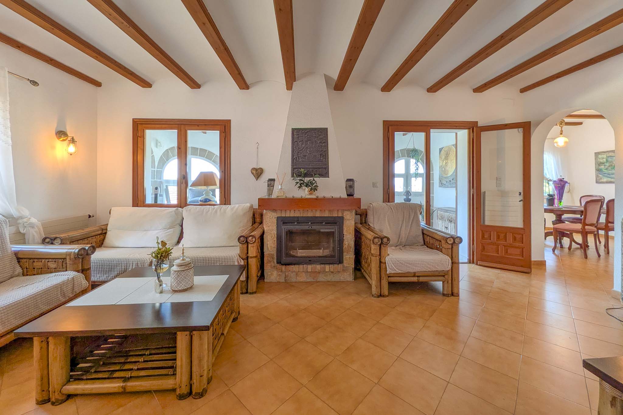 Salón rústico con chimenea de piedra, vigas de madera vista y suelos de gres. El mobiliario de bambú y el acceso al comedor realzan su marcado carácter mediterráneo.