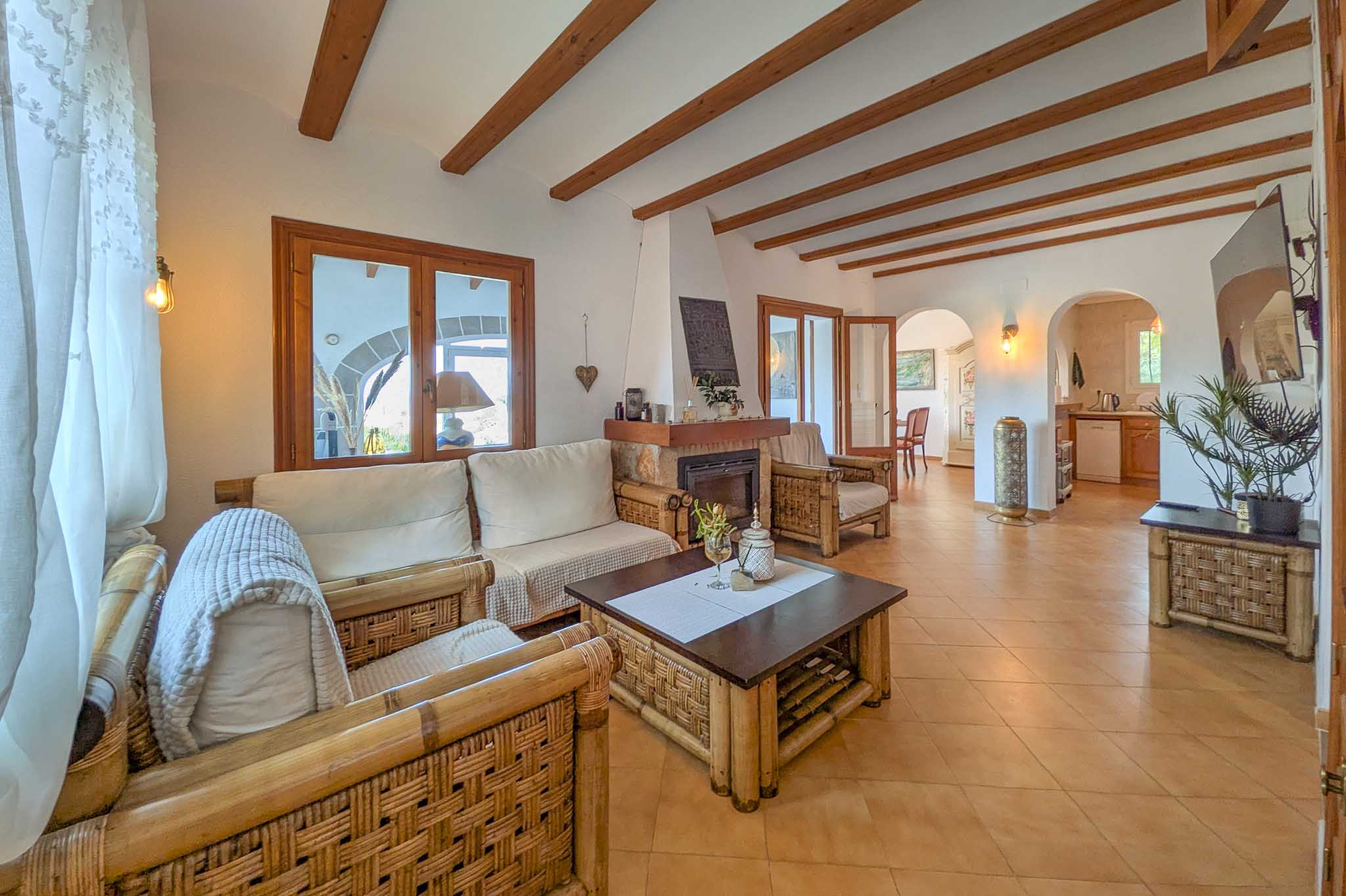 Salón rústico con vigas de madera vistas, chimenea de piedra y mobiliario de bambú. El espacio conecta mediante arcos con el comedor y la cocina.