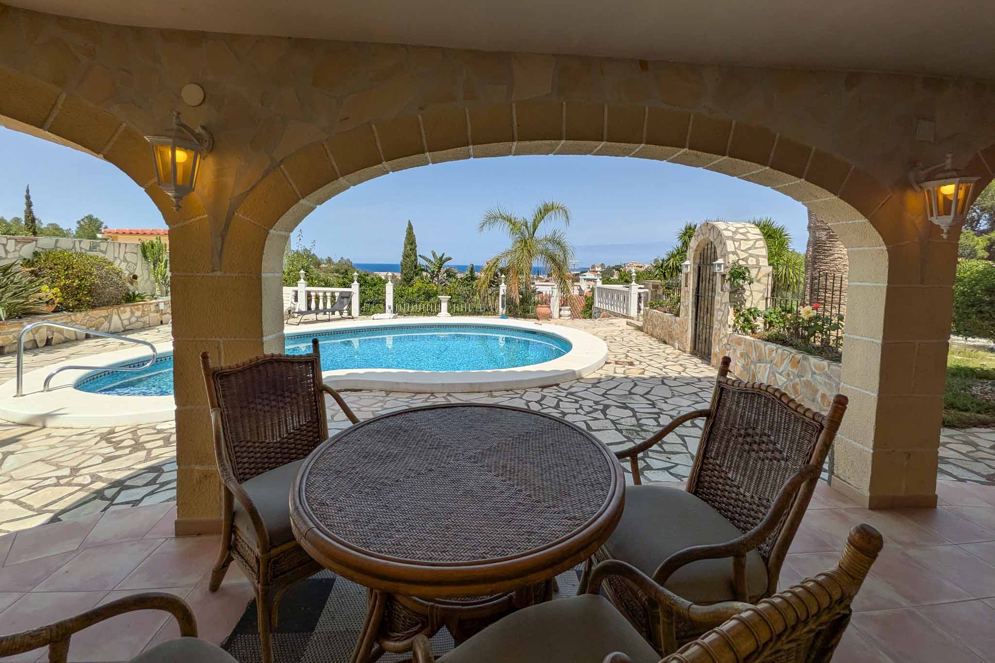 Terraza cubierta con arcos de piedra y mobiliario de mimbre, con vistas directas a la piscina y al mar Mediterráneo en el horizonte.