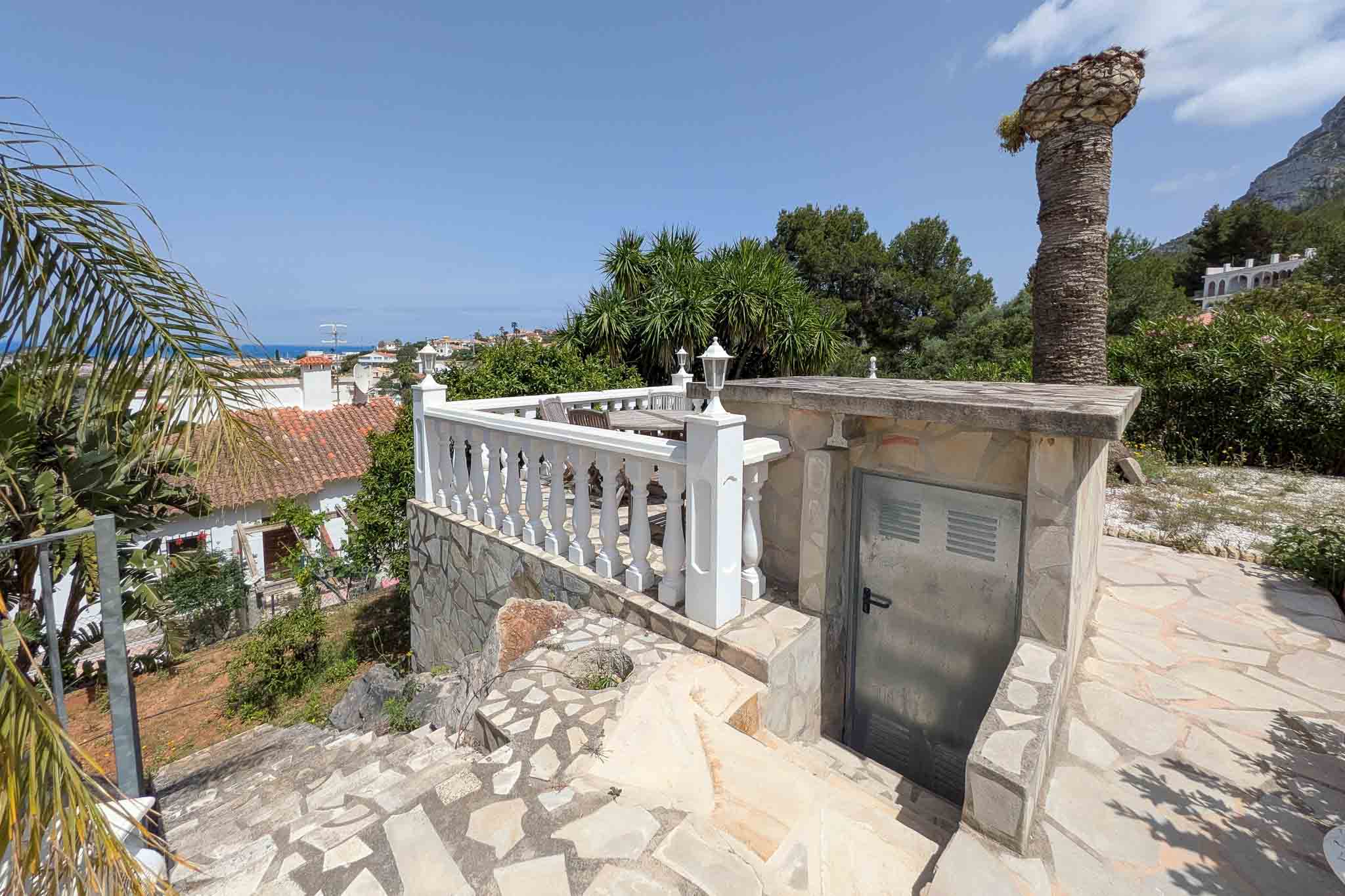 Terraza exterior con balaustrada blanca y suelos de piedra natural, ofreciendo vistas despejadas al mar y acceso a una zona de almacenamiento técnico.