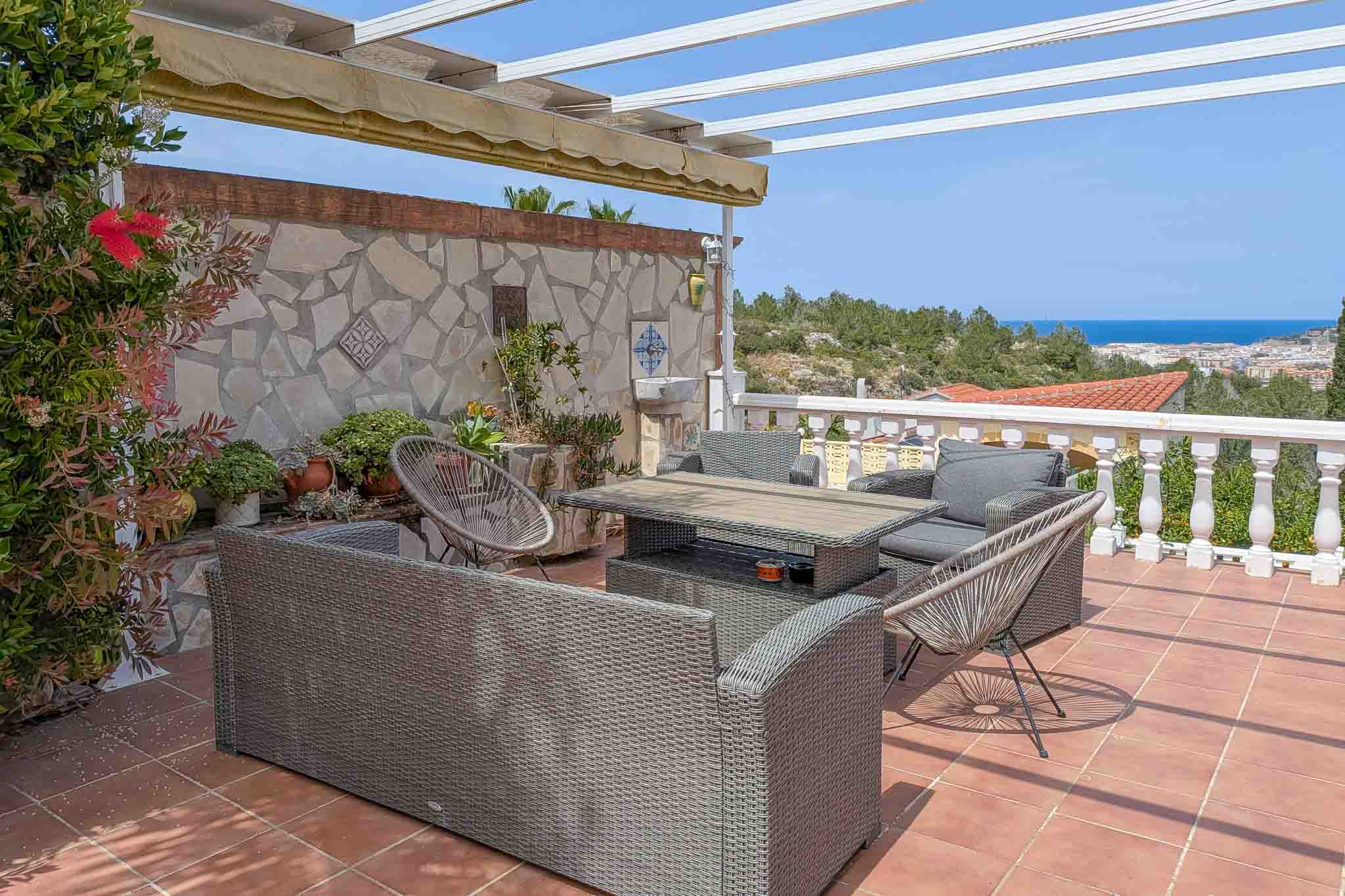 Terraza exterior con mobiliario de ratán, suelo de terracota y vistas panorámicas al mar y Dénia. Incluye pérgola con toldo y pared de piedra natural decorativa.