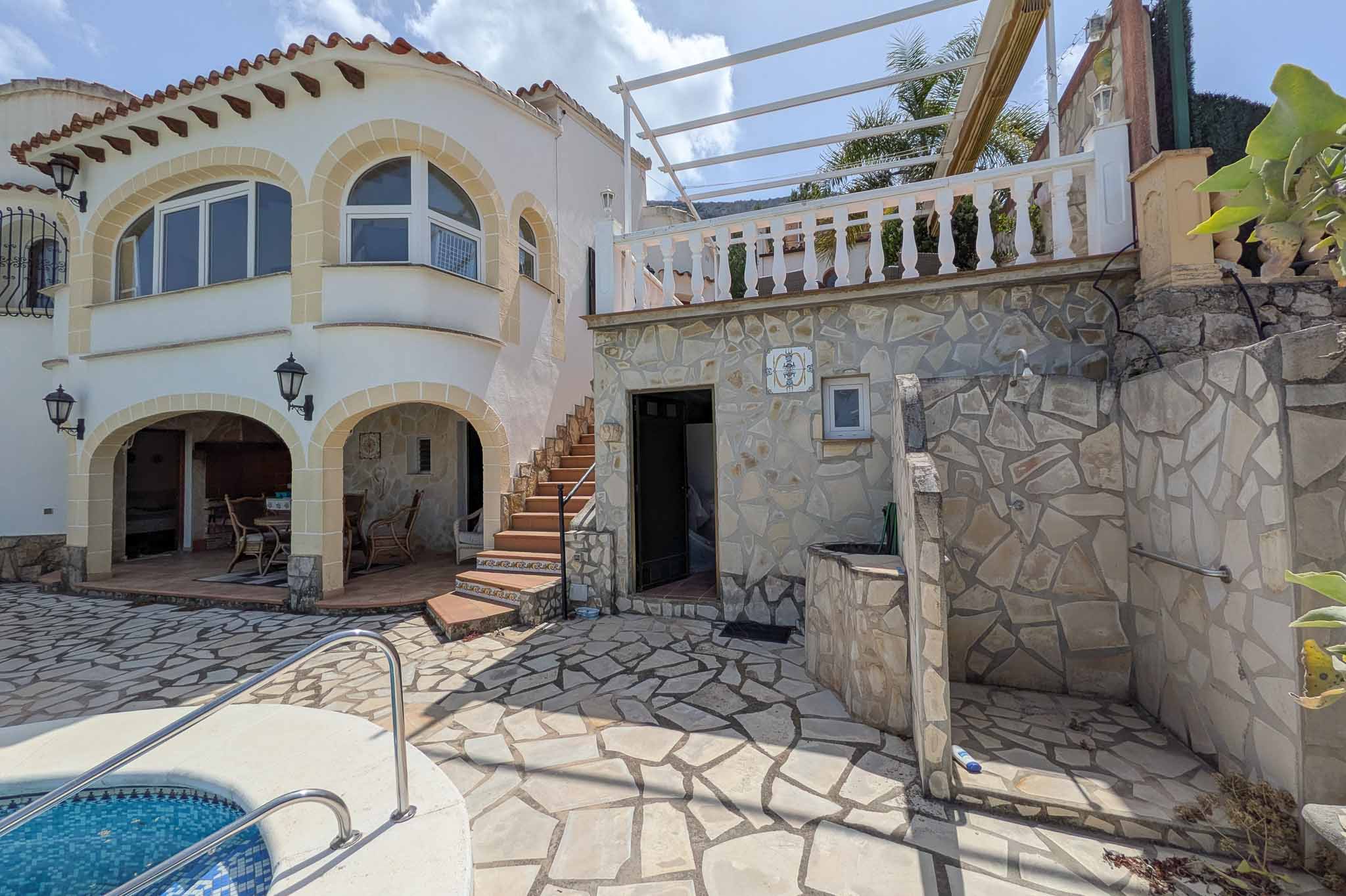 Terraza exterior con suelo de piedra natural, zona de ducha integrada y escaleras hacia el nivel superior con balaustrada blanca y pérgola.
