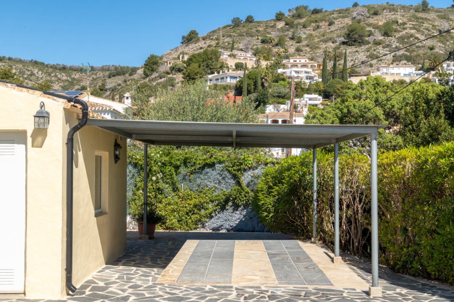 Área de aparcamiento cubierto tipo carport con estructura metálica y suelo de piedra natural, situada junto a la fachada y rodeada de vegetación.