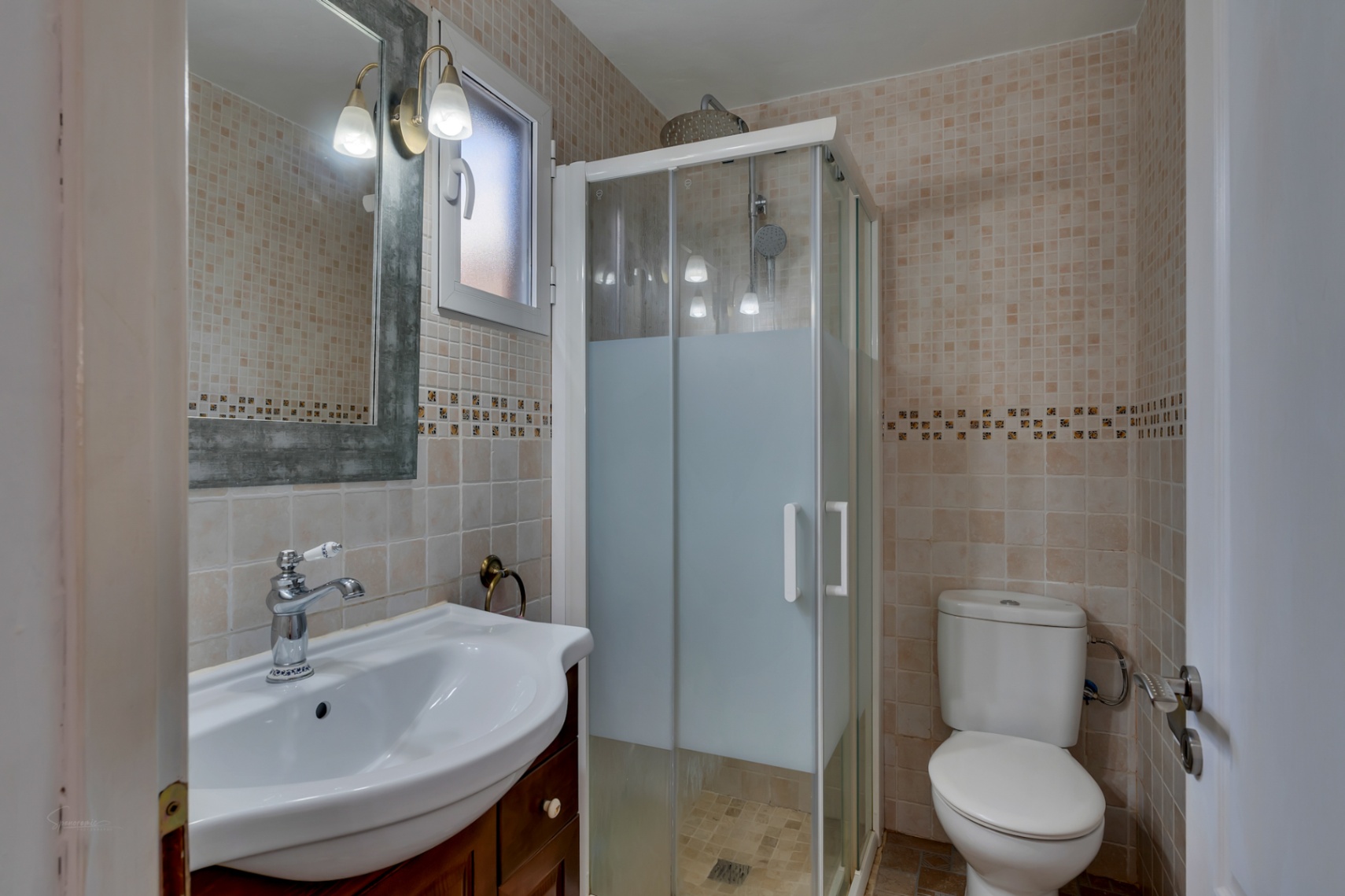 Baño funcional con paredes alicatadas en tonos beige, plato de ducha con mampara translúcida, lavabo sobre mueble de madera y espejo con apliques clásicos.