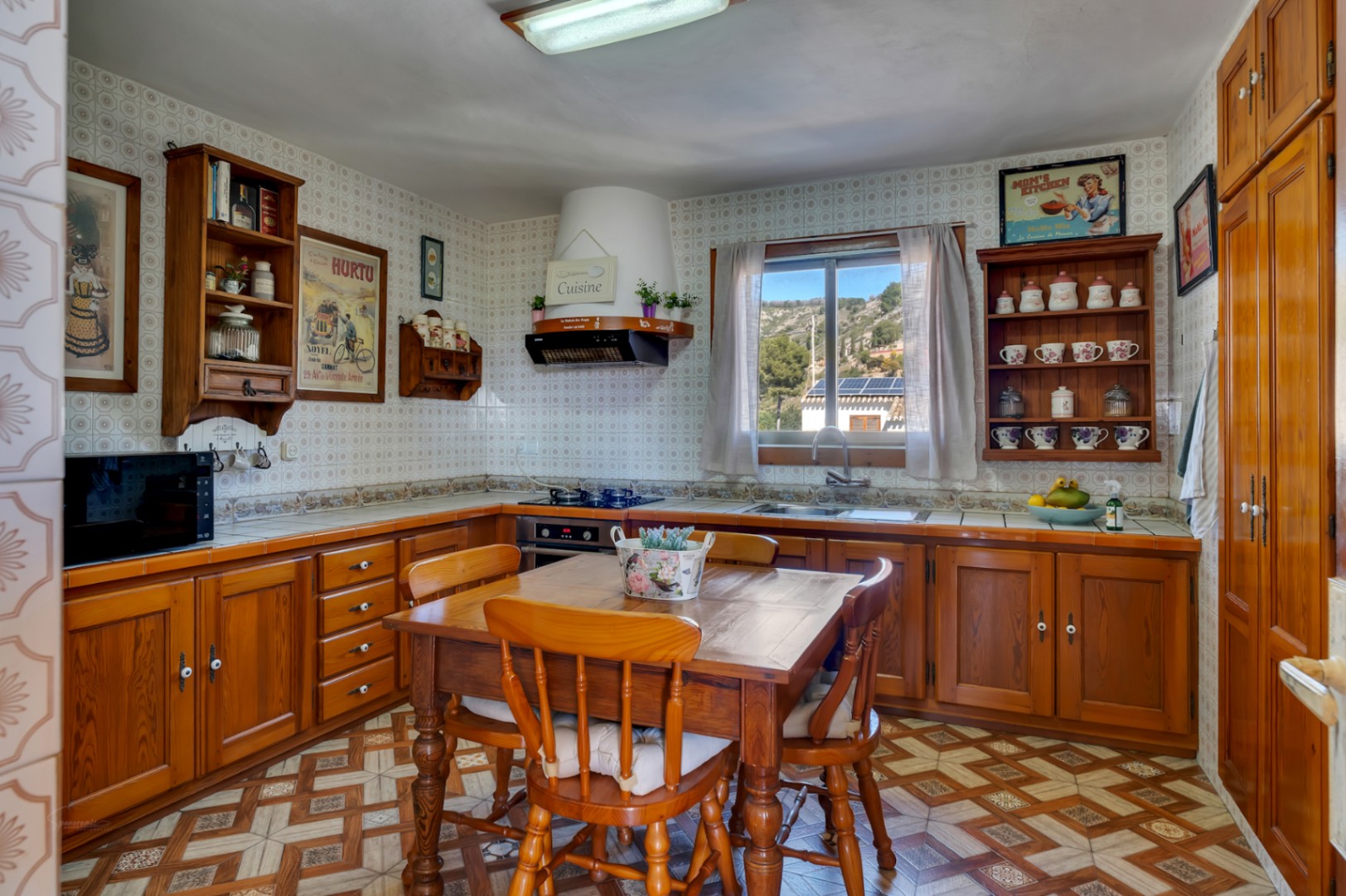 Cocina de estilo rústico con armarios de madera, mesa de comedor central y azulejos decorativos. Dispone de luz natural y vistas despejadas a través de su ventana.