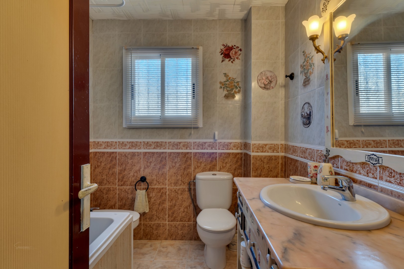 Cuarto de baño de estilo clásico con encimera de mármol, lavabo ovalado, sanitarios blancos y azulejos decorados con motivos florales y cenefas.