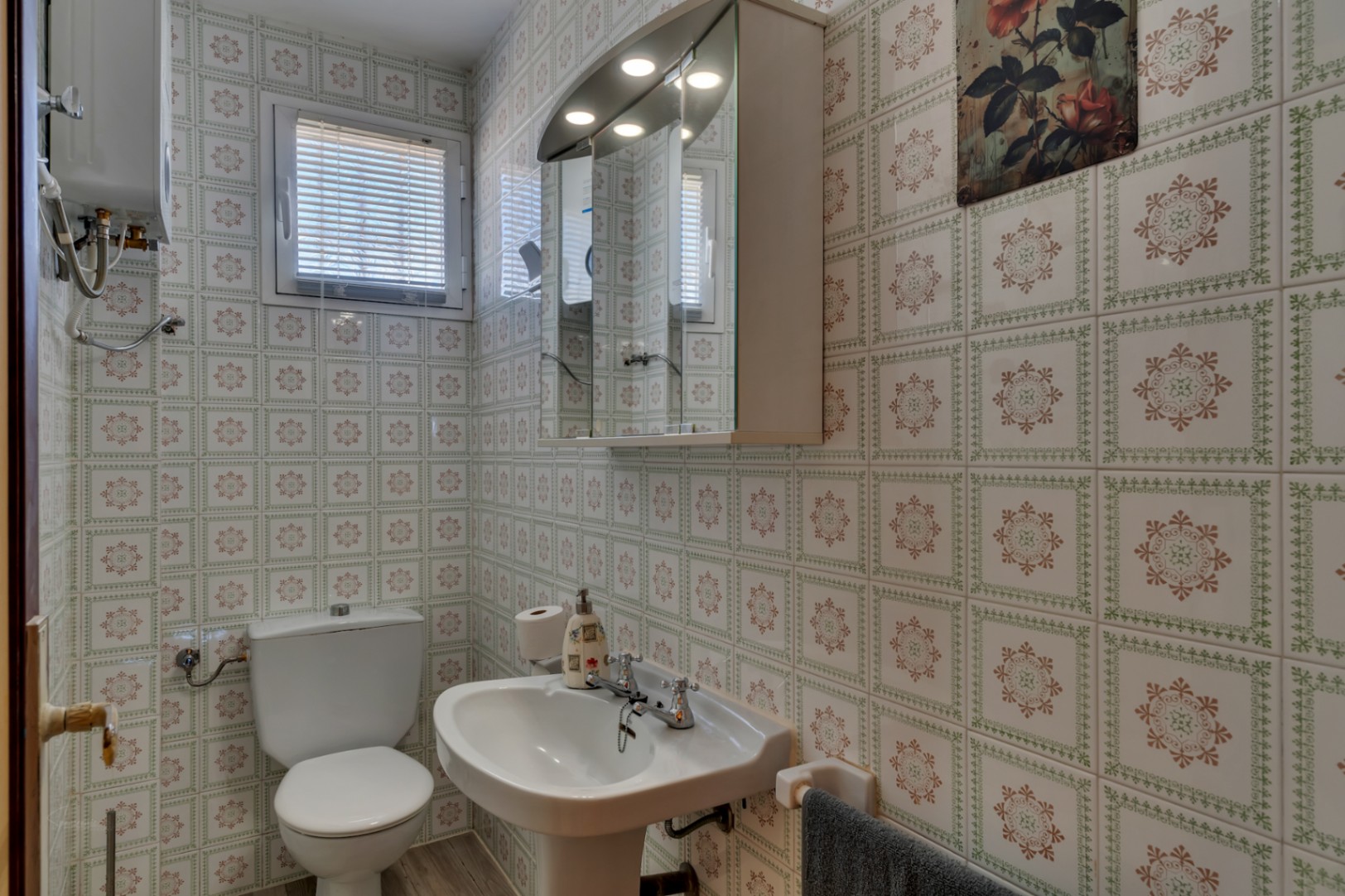 Cuarto de baño funcional con paredes alicatadas con motivos geométricos, lavabo de pedestal, inodoro y armario de espejo con iluminación integrada.