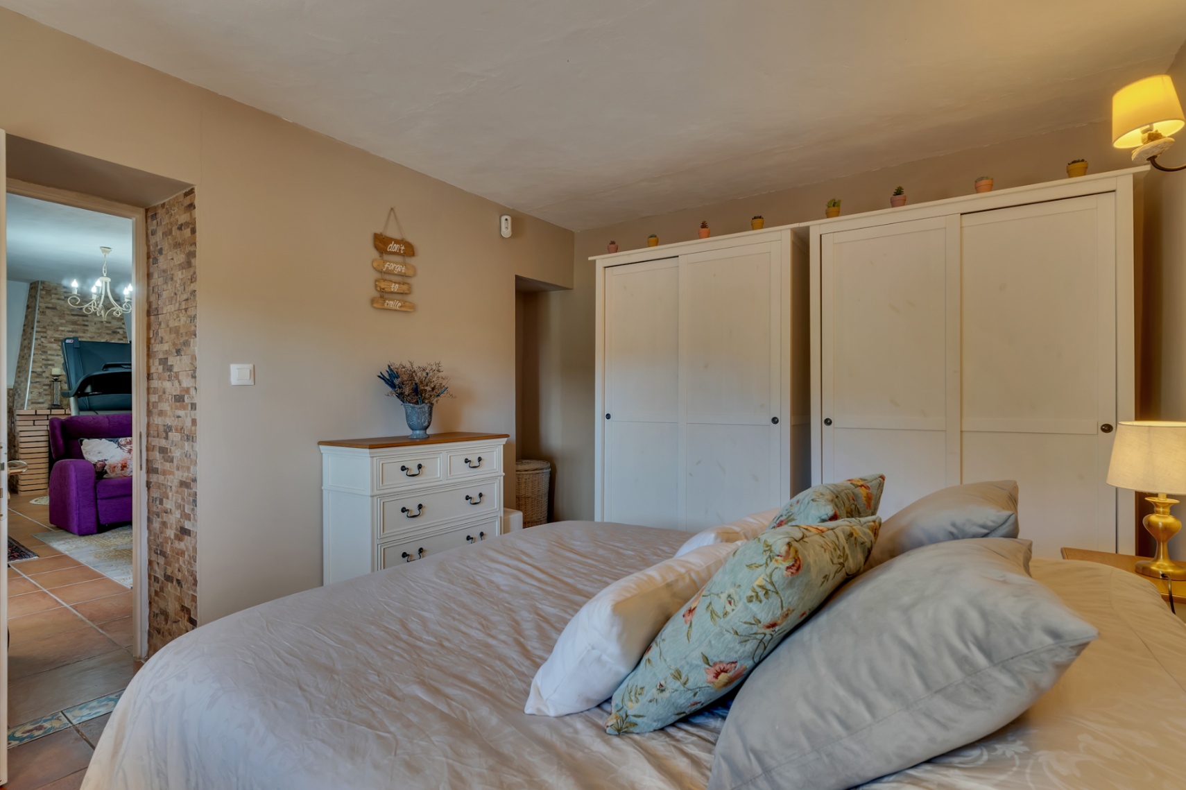 Dormitorio acogedor con armarios blancos, cómoda de madera y paredes en tonos tierra. Destaca el detalle de piedra natural en el arco que conecta con la zona de estar.