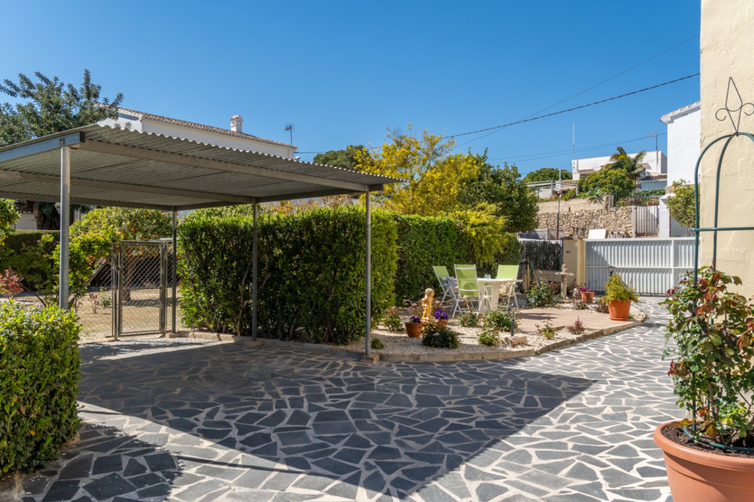 Entrada a la propiedad con suelo de piedra irregular, carport metálico y zona ajardinada con setos y plantas ornamentales bajo el sol mediterráneo.