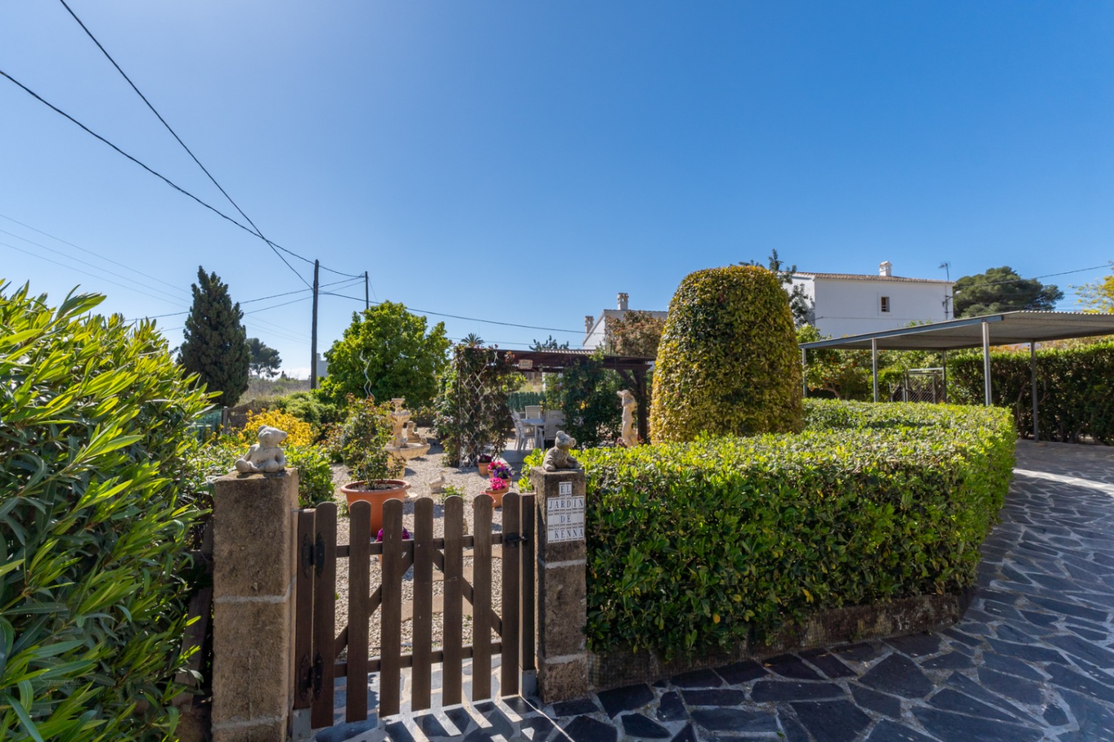 Entrada al jardín mediterráneo con puerta de madera, setos cuidados, zona de grava y una pérgola sombreada junto a un camino empedrado.