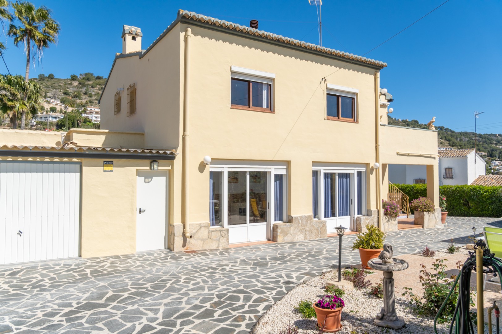 Exterior de la villa con fachada en tonos crema, zócalo de piedra natural y amplio patio pavimentado. Incluye garaje cerrado y ventanales blancos en la planta baja.