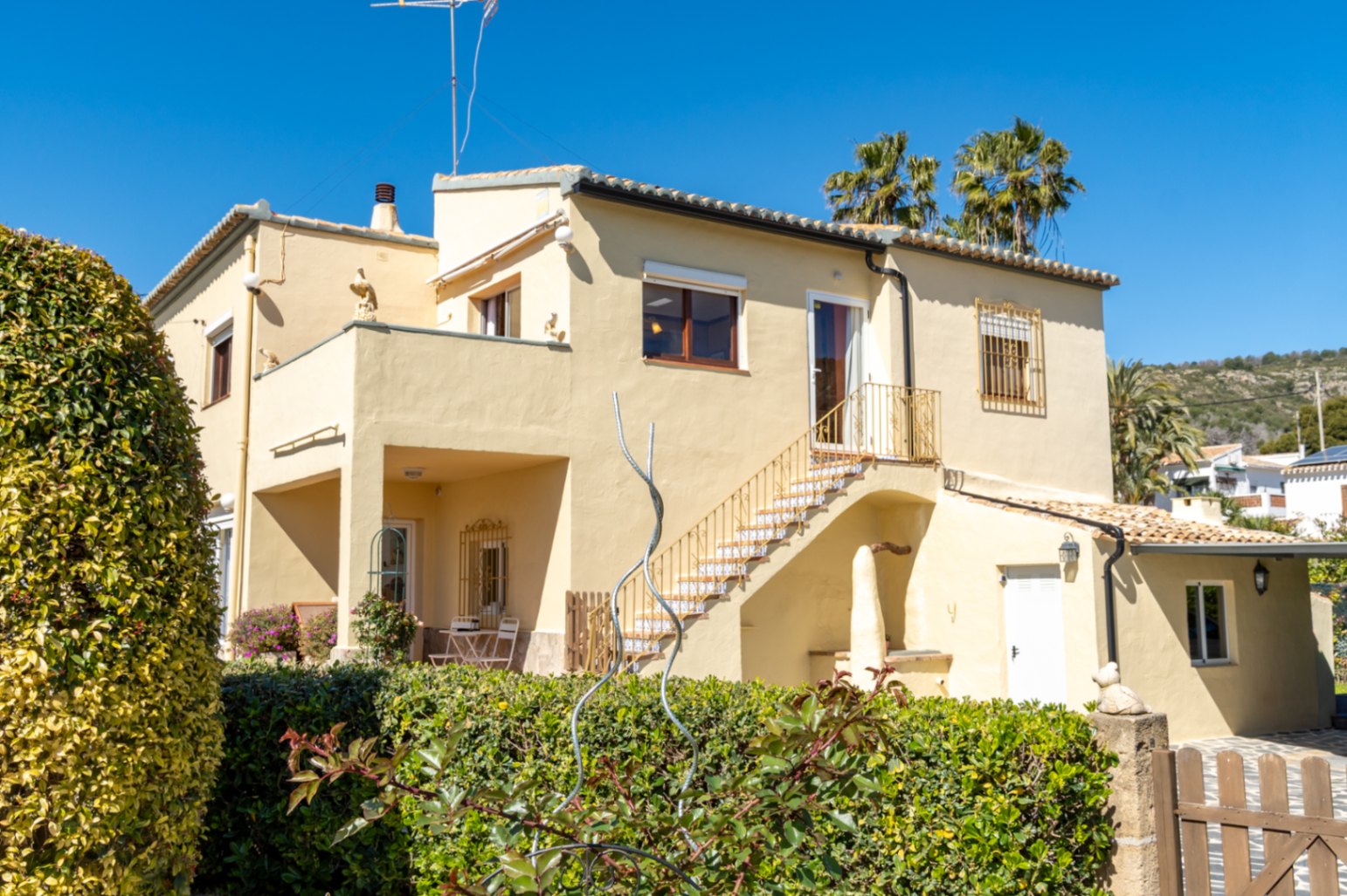 Fachada exterior de la villa con escalera lateral de azulejos, terrazas en dos niveles y setos perimetrales bajo un cielo despejado en Jávea.