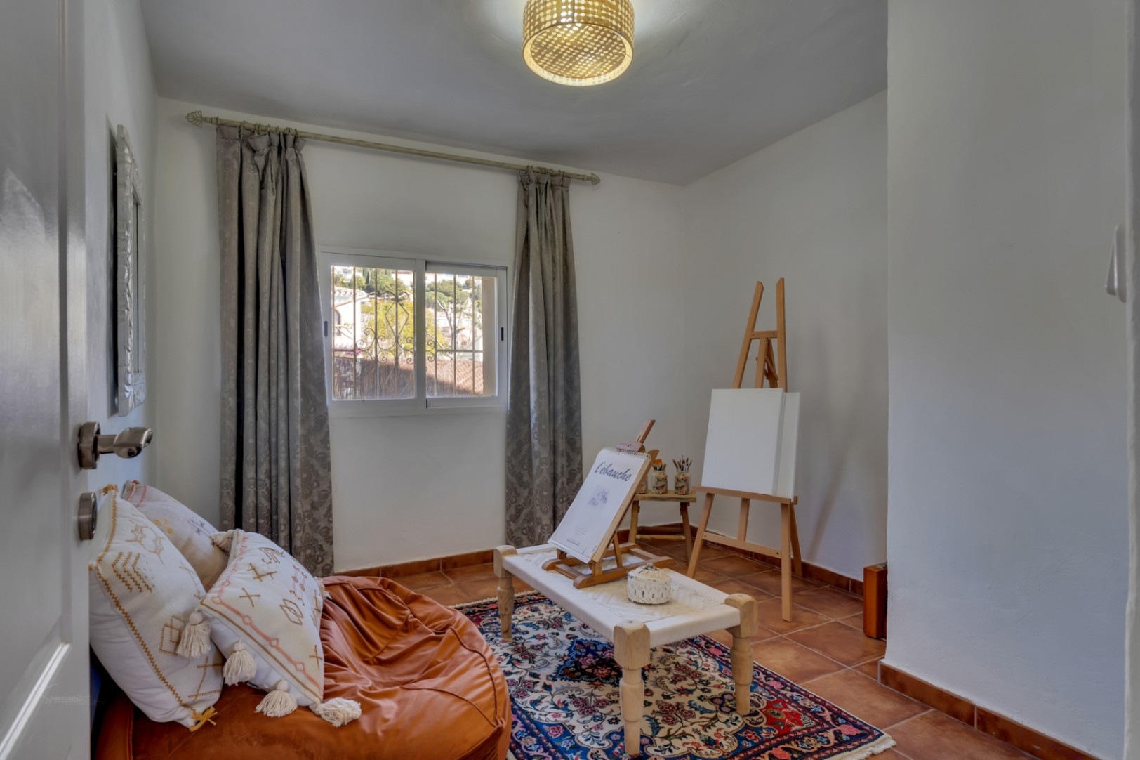 Habitación versátil configurada como estudio de arte con suelos de terracota, caballete de madera, luz natural y una decoración acogedora de estilo mediterráneo.