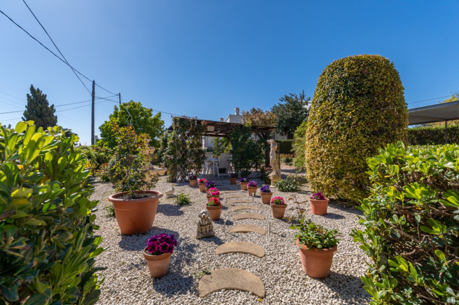 Jardín mediterráneo con suelo de grava blanca, camino de piedras y macetas de terracota. Incluye una pérgola de madera con zona de comedor exterior bajo el sol.
