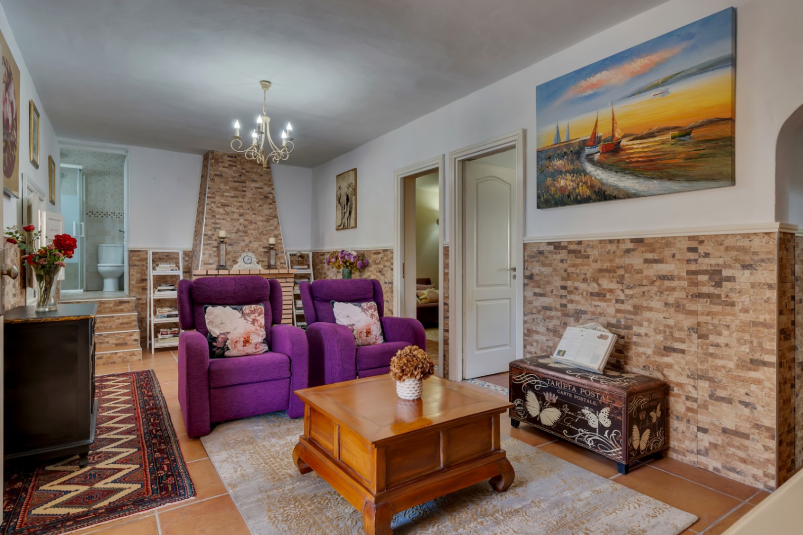 Salón acogedor con chimenea revestida en piedra, butacas color púrpura y acceso directo a un baño completo, destacando las paredes con zócalo cerámico.