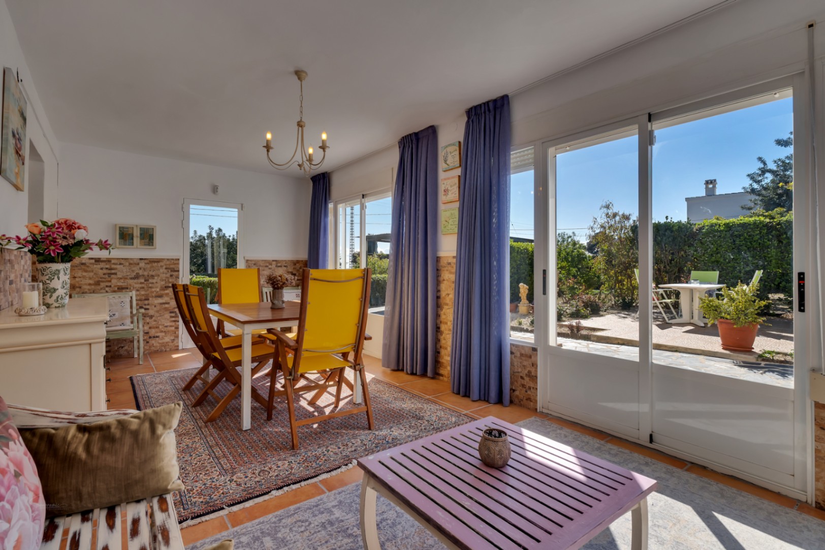 Salón comedor luminoso con zócalo de piedra, mobiliario en tonos amarillos y grandes ventanales correderos que conectan con la terraza exterior y el jardín.